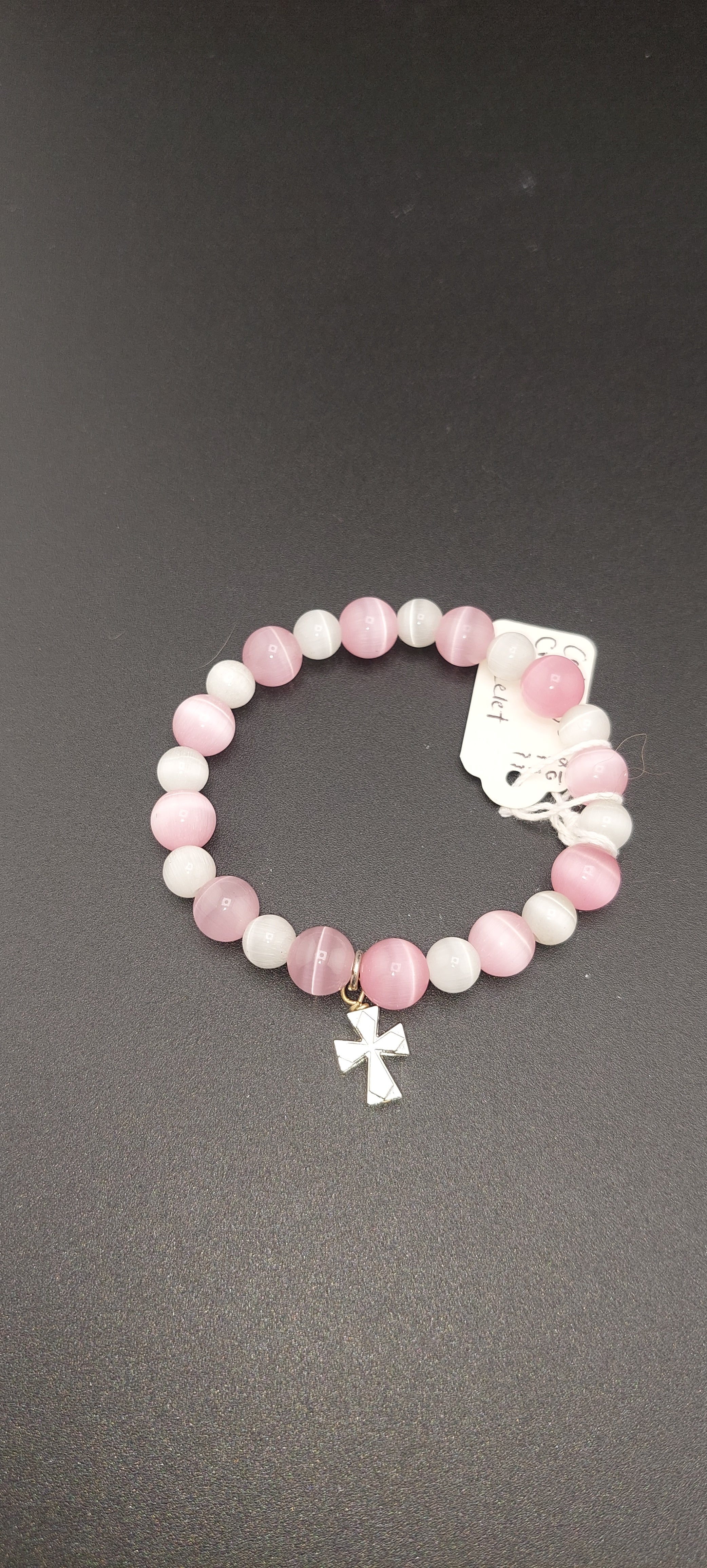 A pink cat eye cross bracelet with white and pink beads, and a silver cross charm, displayed against a black background.