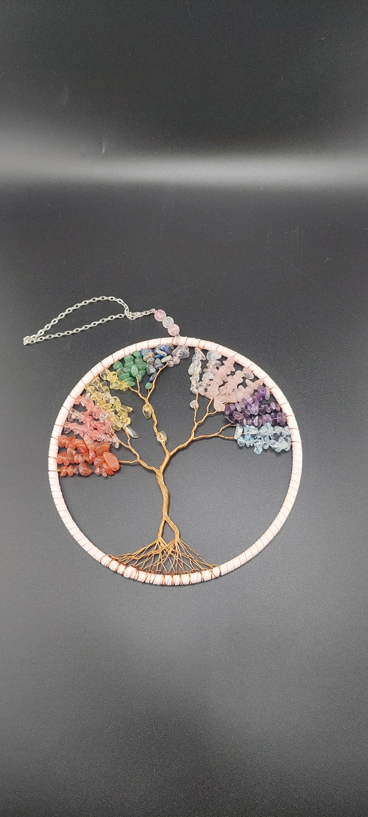 A dream catcher featuring a tree of life design, with multicolored stones arranged along its branches and roots, set against a black background.