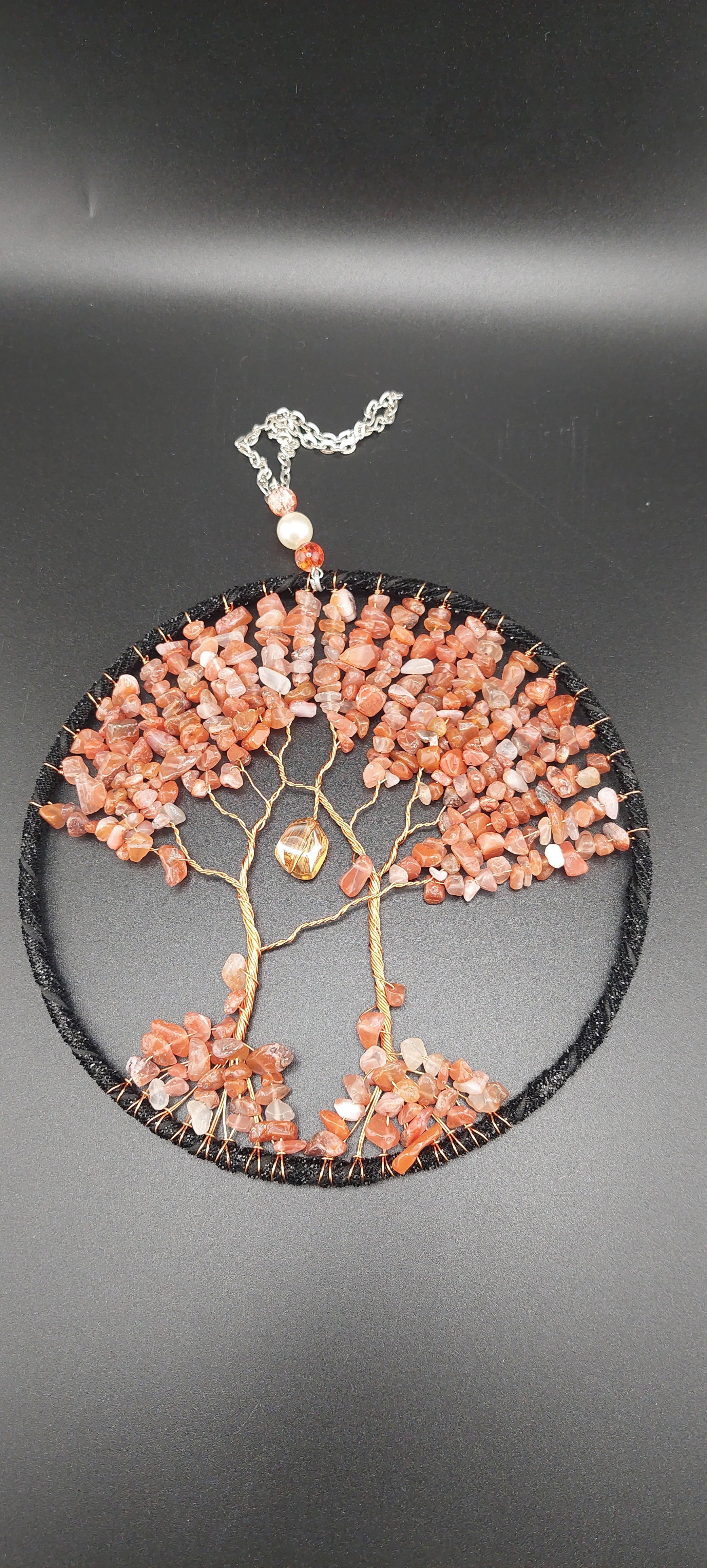A dream catcher featuring a web-like pattern with carnelian stones, predominantly in shades of orange and white, with a small feather at the bottom.