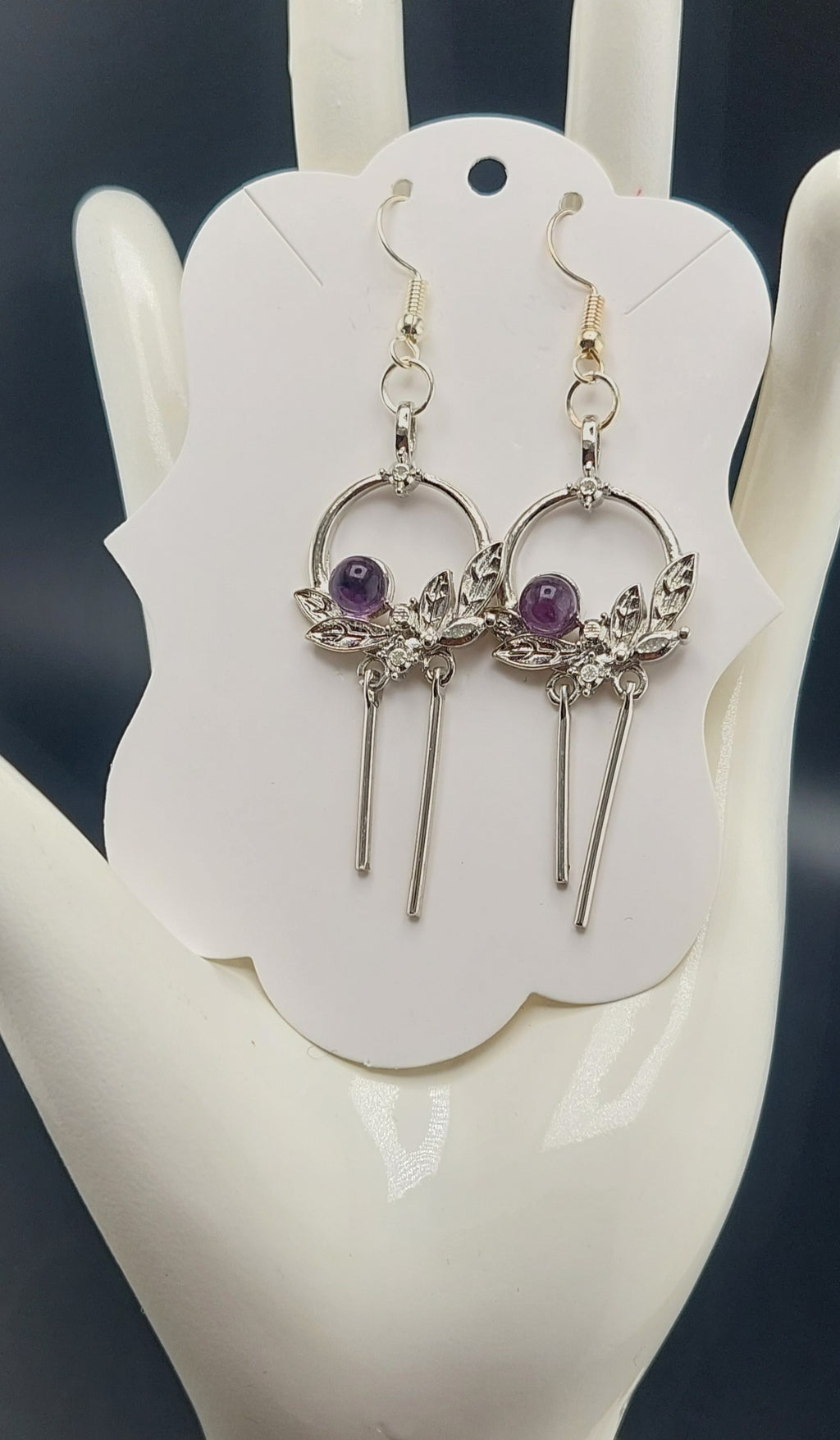 A pair of silver-colored flower wreath earrings with purple stones, displayed on a white card.