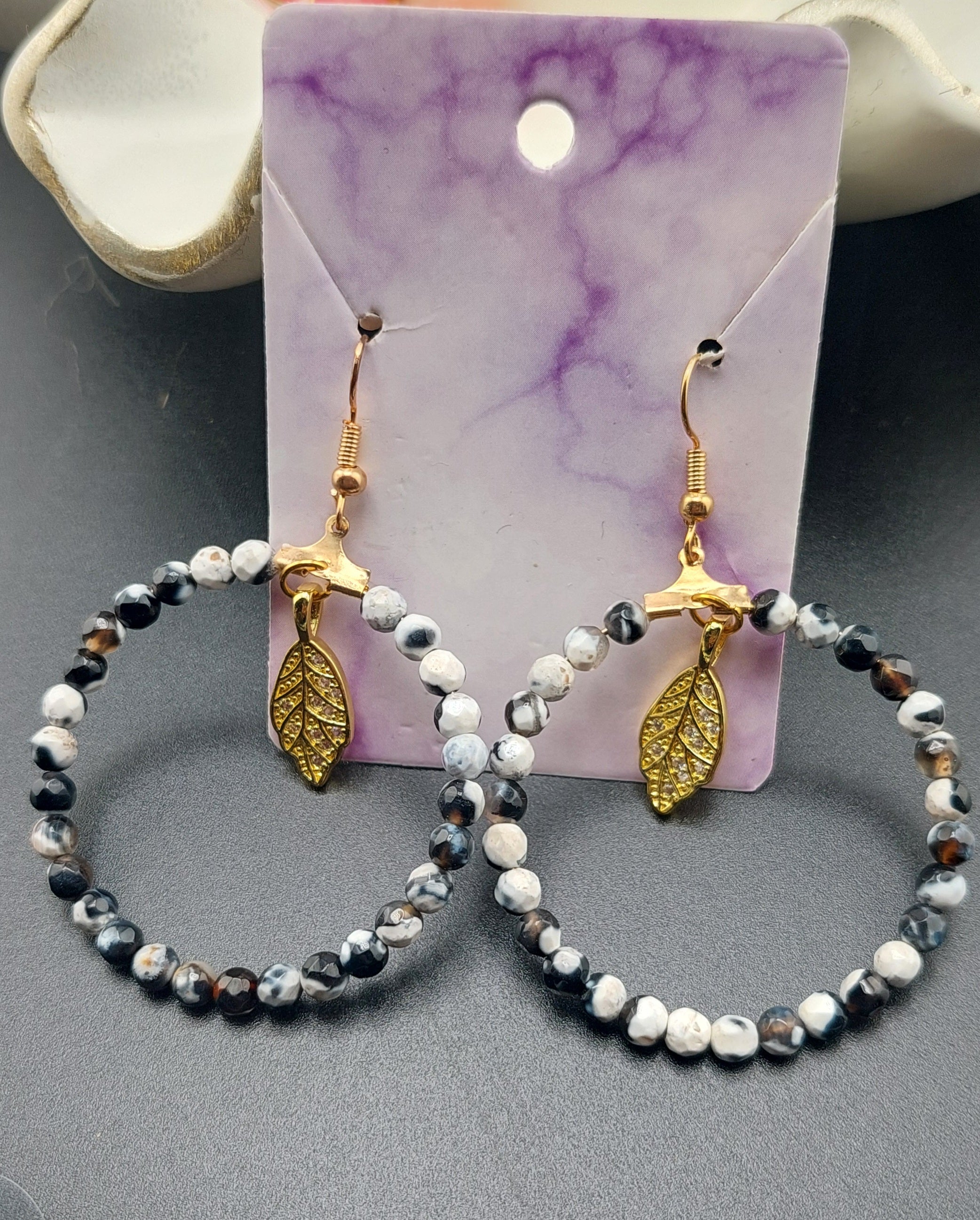 A pair of black and white beaded earrings with gold-colored leaf charms, displayed on a pink card.