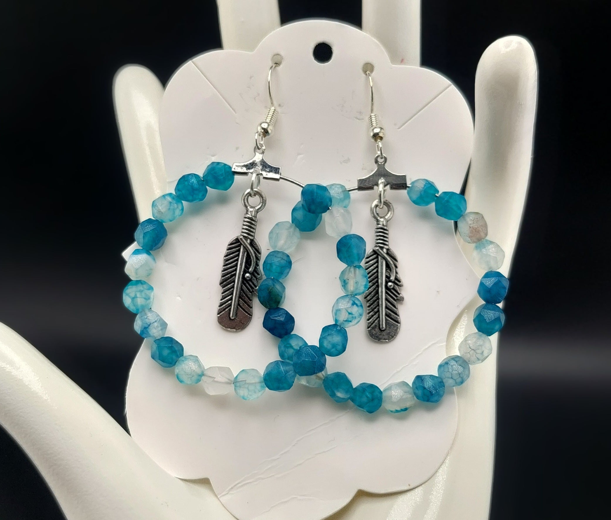A pair of blue lace agate hoop earrings with feather accents displayed on a white stand.