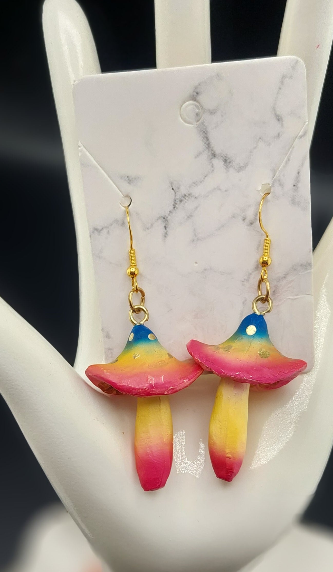 A pair of rainbow-colored mushroom earrings displayed on a white card against a marble background.