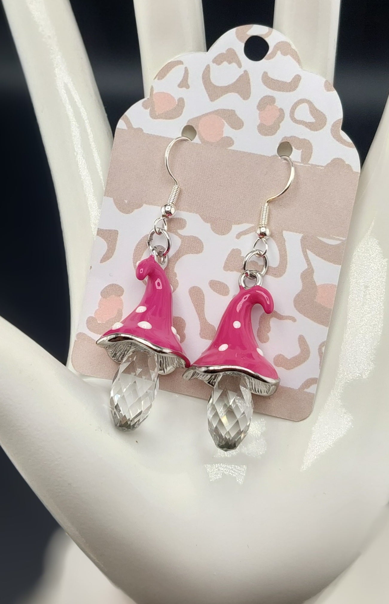 A pair of pink mushroom-shaped earrings with white dots and clear crystal-like pendants, displayed on a beige card with a leopard print design.