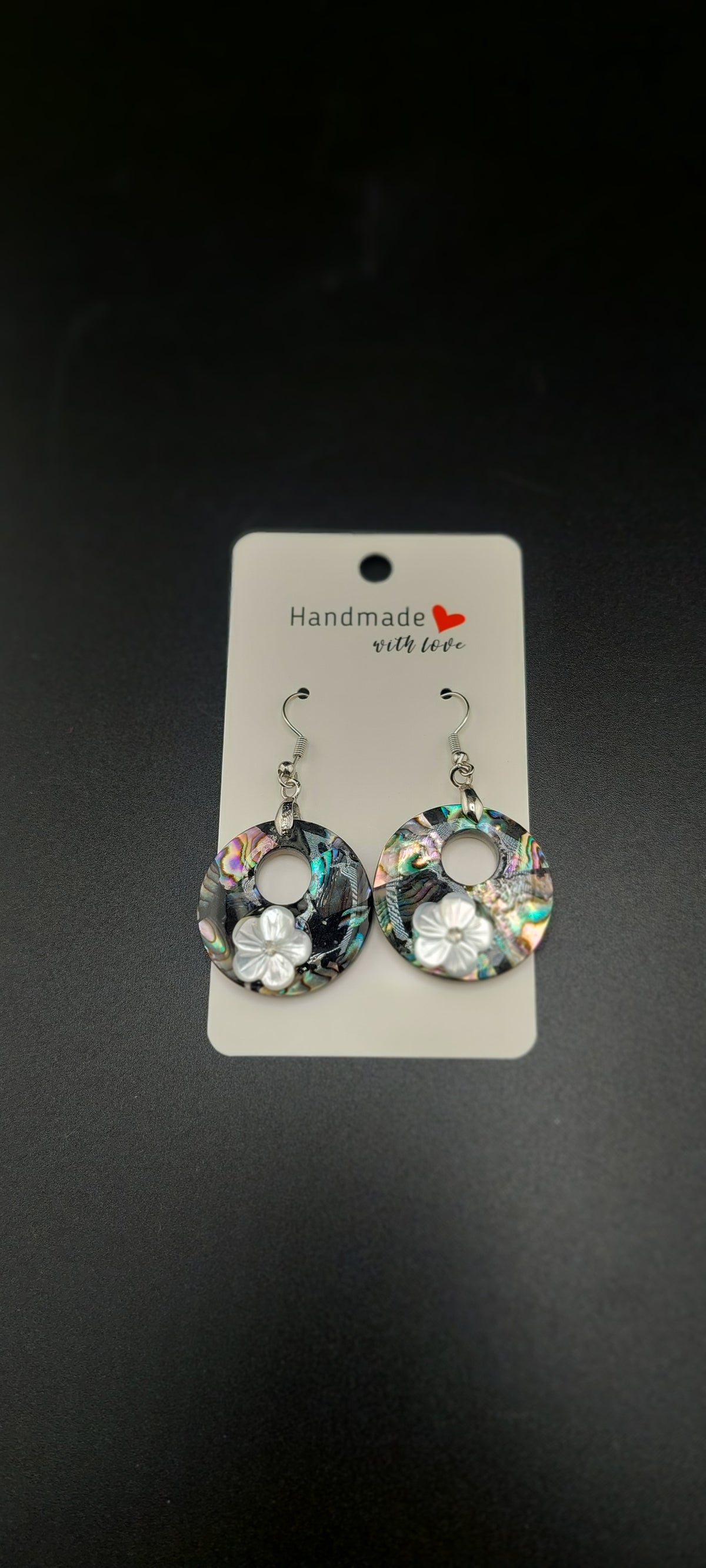 A pair of shell-shaped earrings with flower patterns, displayed on a cardboard holder against a black background.