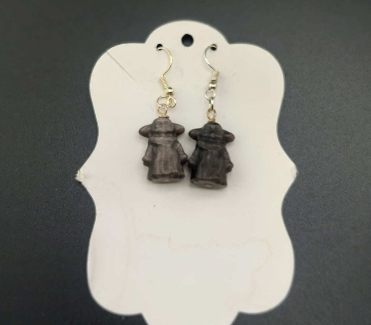 A pair of Yoda-shaped earrings displayed on a white holder, with a dark stone material and silver wire.