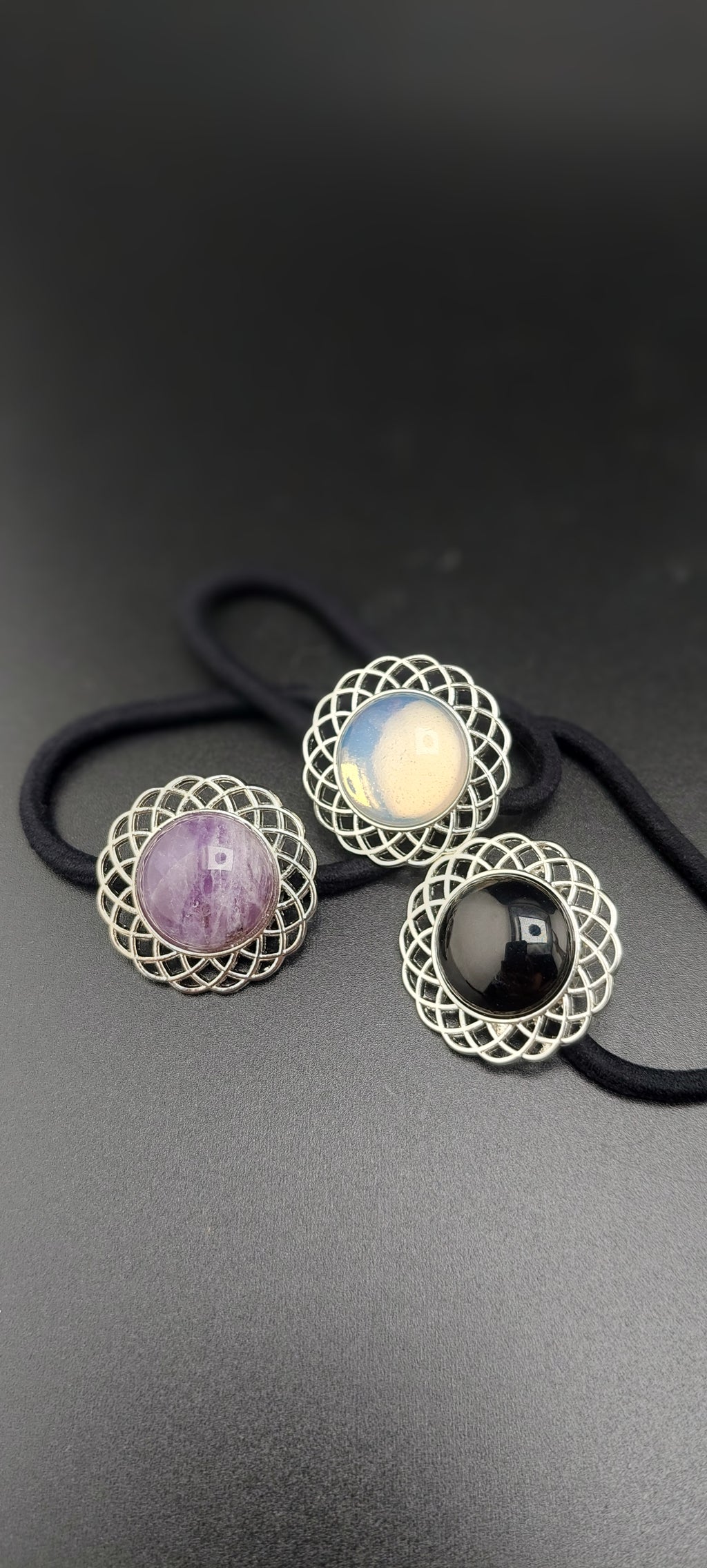 A set of three silver hair ties with different healing stones attached to them, displayed against a dark background.