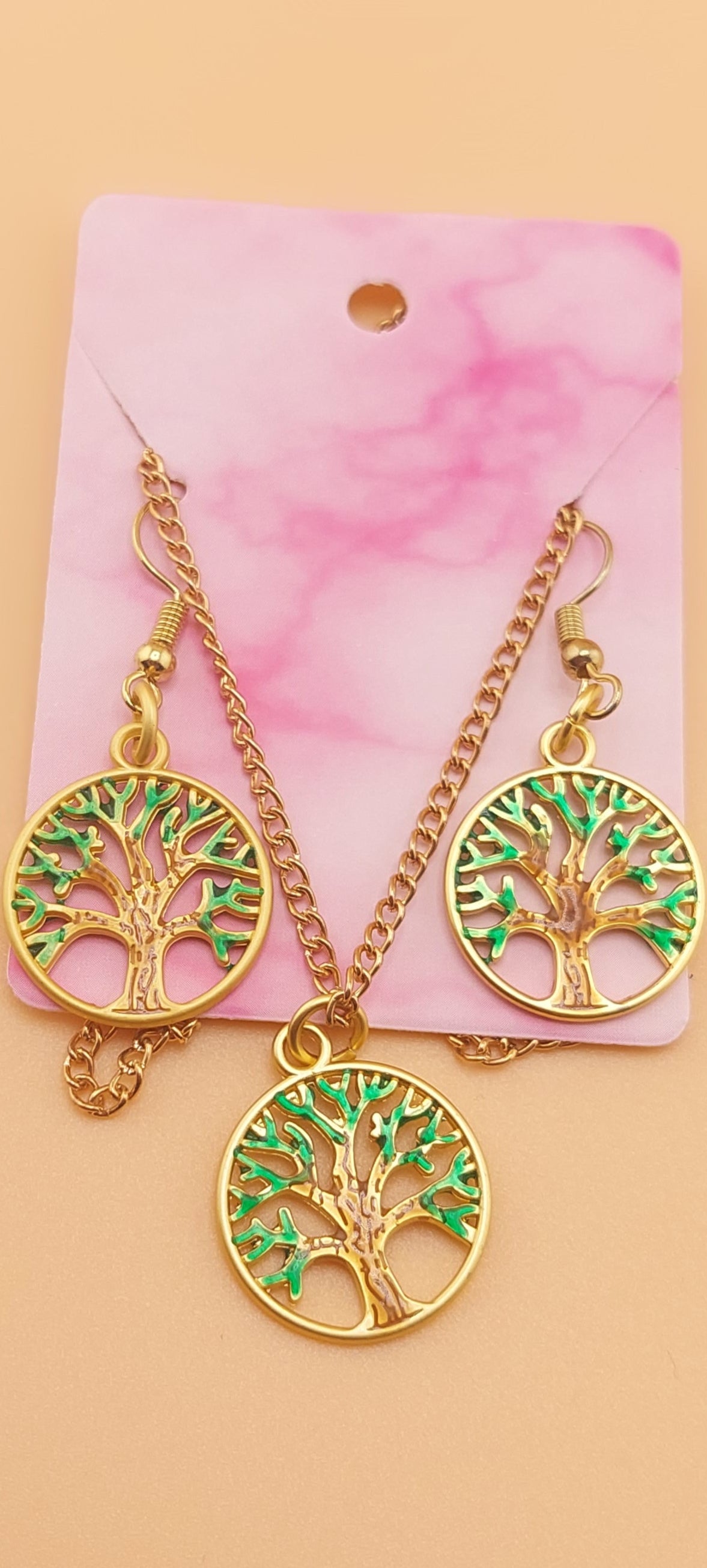 A gold-colored metal necklace and earring set featuring a tree of life design, displayed on a pink card.