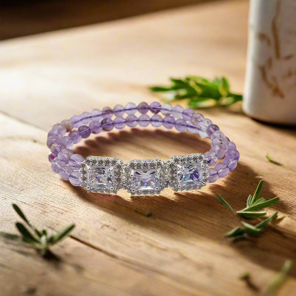 A purple amethyst bracelet with a silver clasp, displayed on a wooden surface with a cup and greenery in the background.