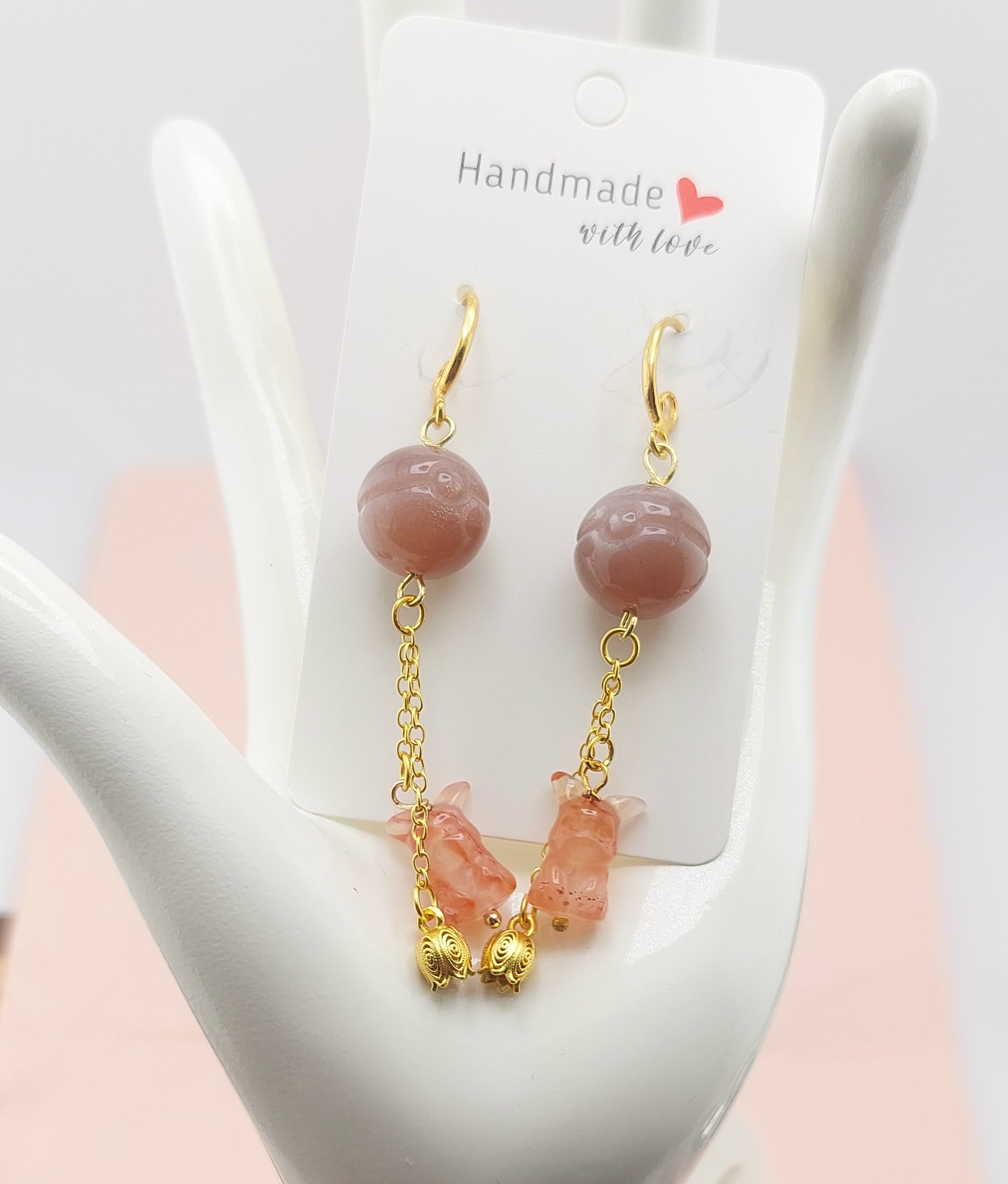 A pair of Pikachu dangle earrings featuring gold-colored wire and pink bead-like elements, displayed on a white stand with 'Handmade with love' text.