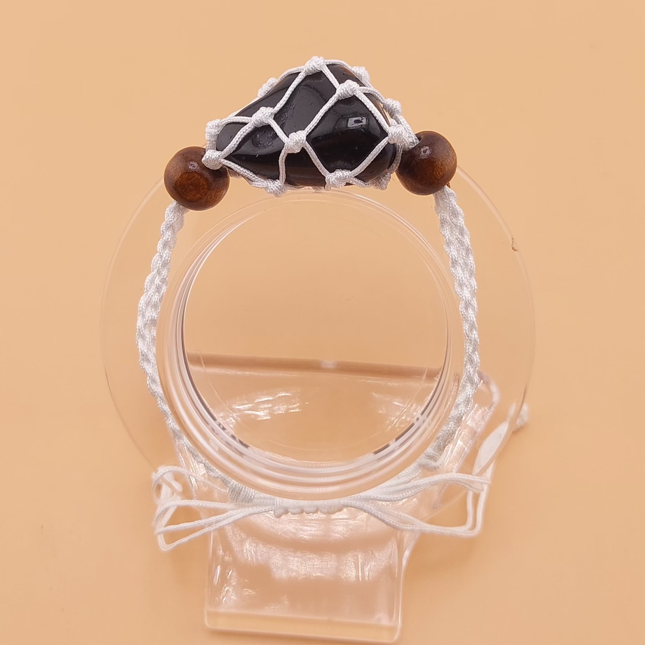 A white Macrame tumbled stone bracelet displayed on a clear stand against a beige background.