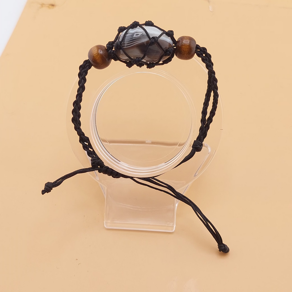 A black Macrame tumbled stone bracelet displayed on a clear stand against a beige background.