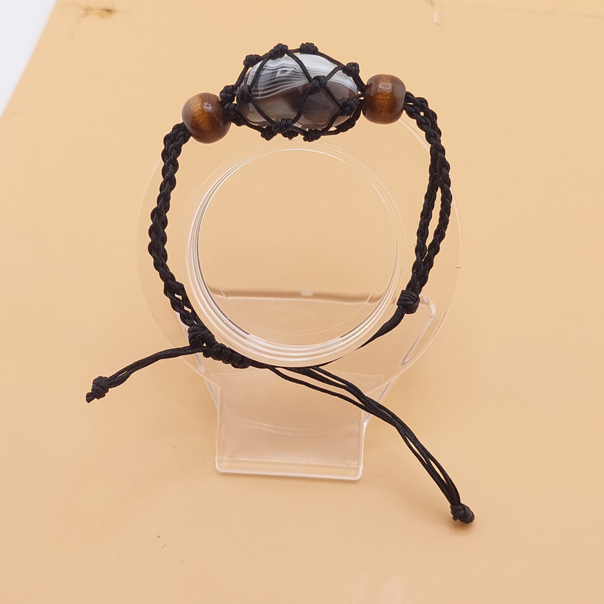 A black Macrame tumbled stone bracelet displayed on a clear stand against a beige background.