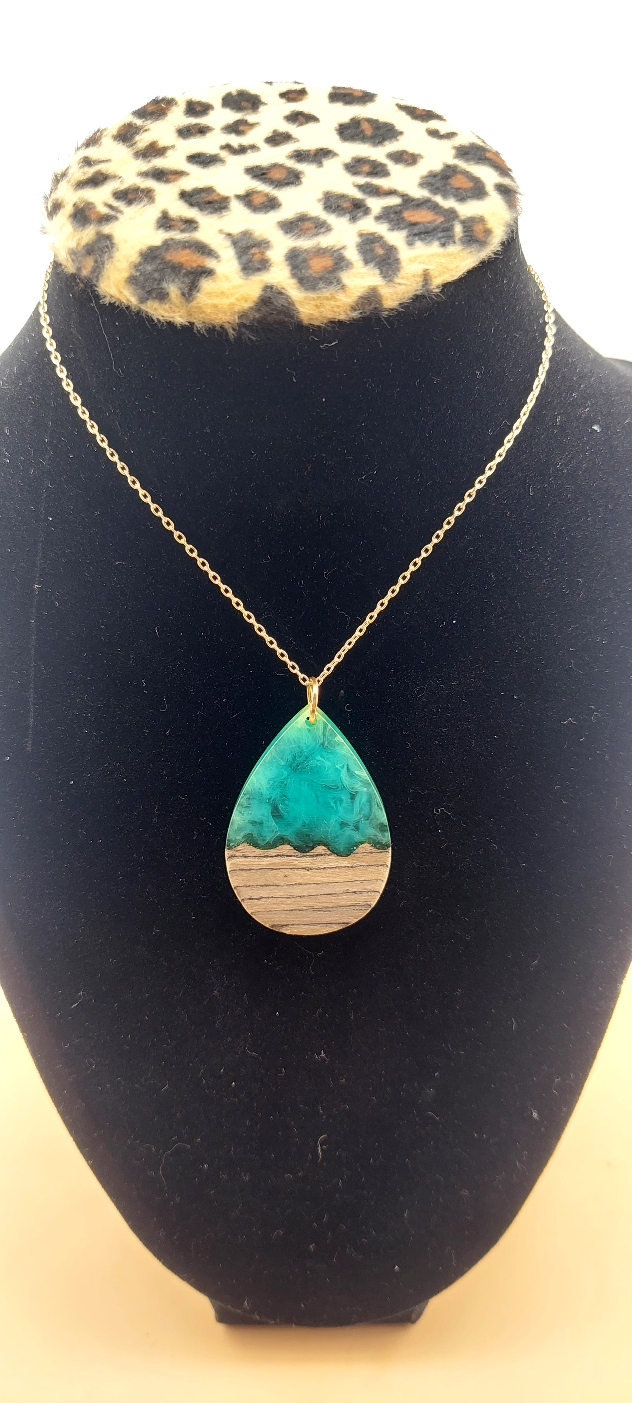 A tear drop-shaped necklace made of resin and wood, displayed on a black mannequin with a leopard print collar.