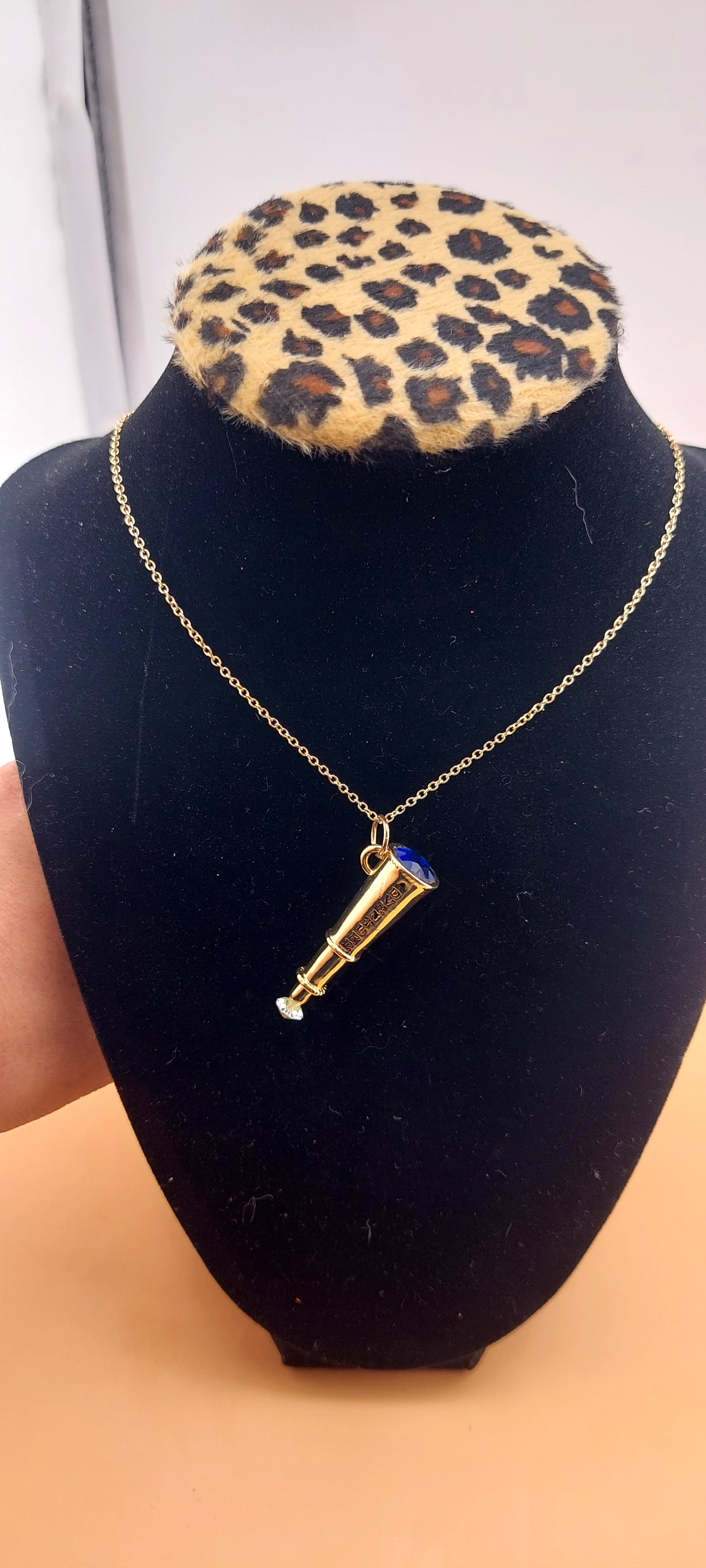 A gold telescope-shaped necklace displayed on a black velvet stand with a leopard print base.