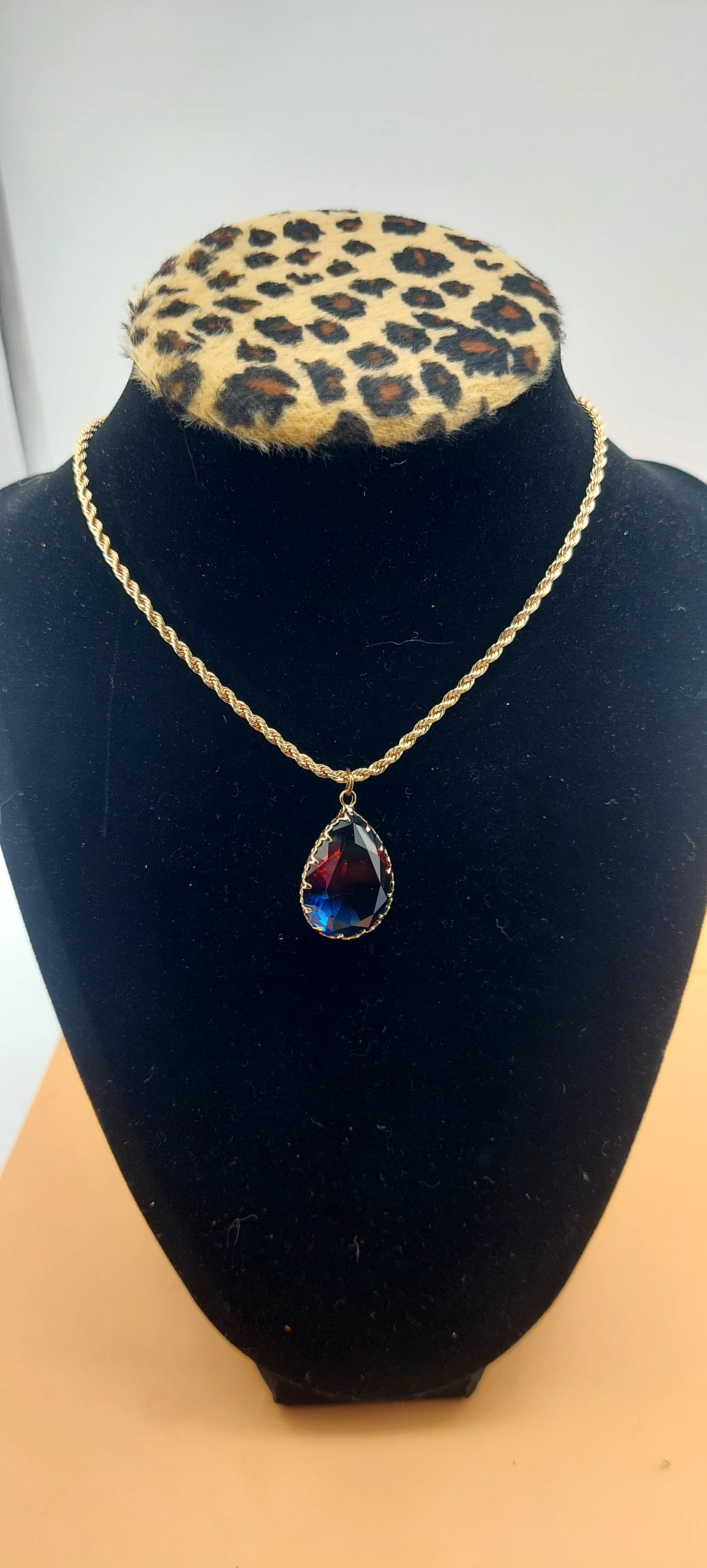A gold necklace with a multicolored gemstone pendant displayed on a black mount with a leopard print pattern.