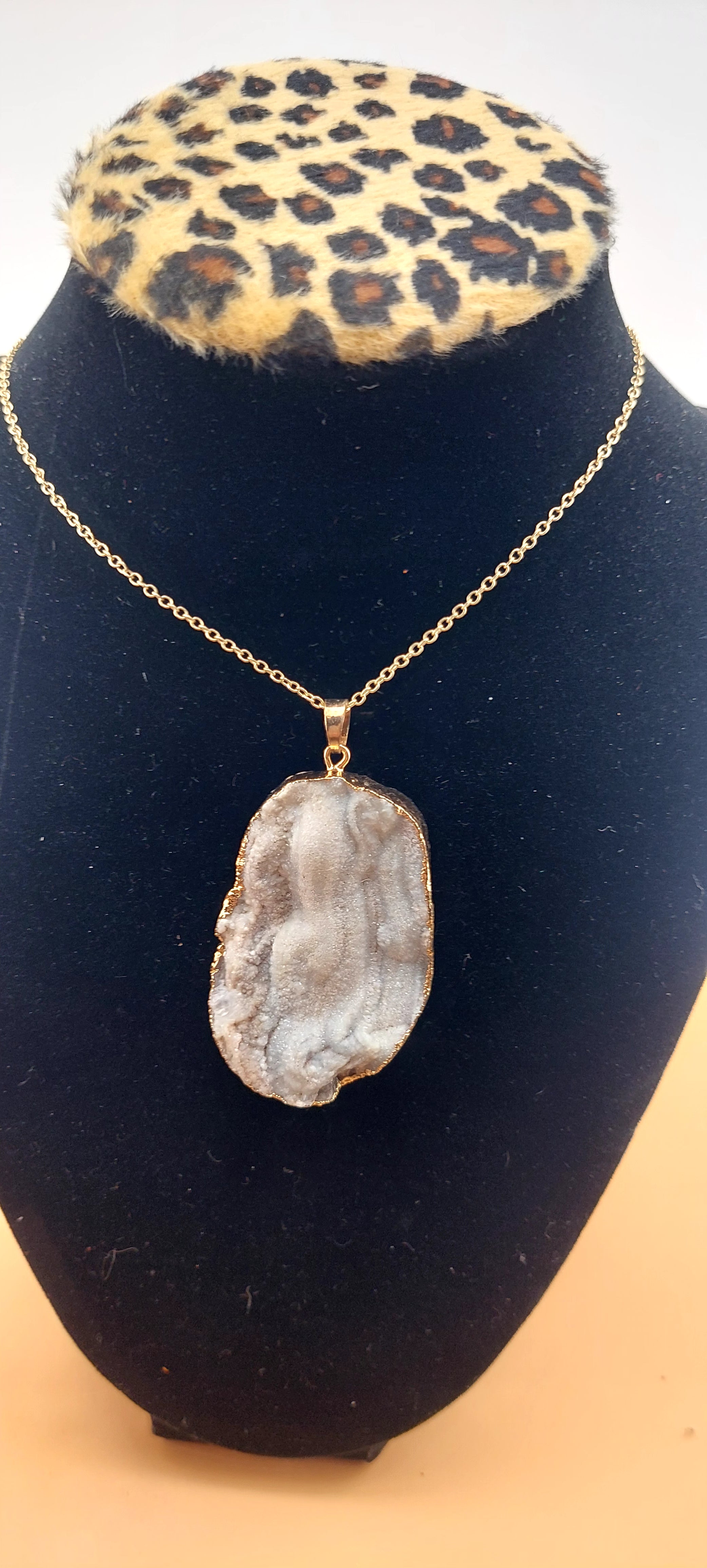 A druzy necklace with a large, irregularly shaped stone pendant mounted on a gold-colored chain, displayed on a black mannequin with a leopard print collar.