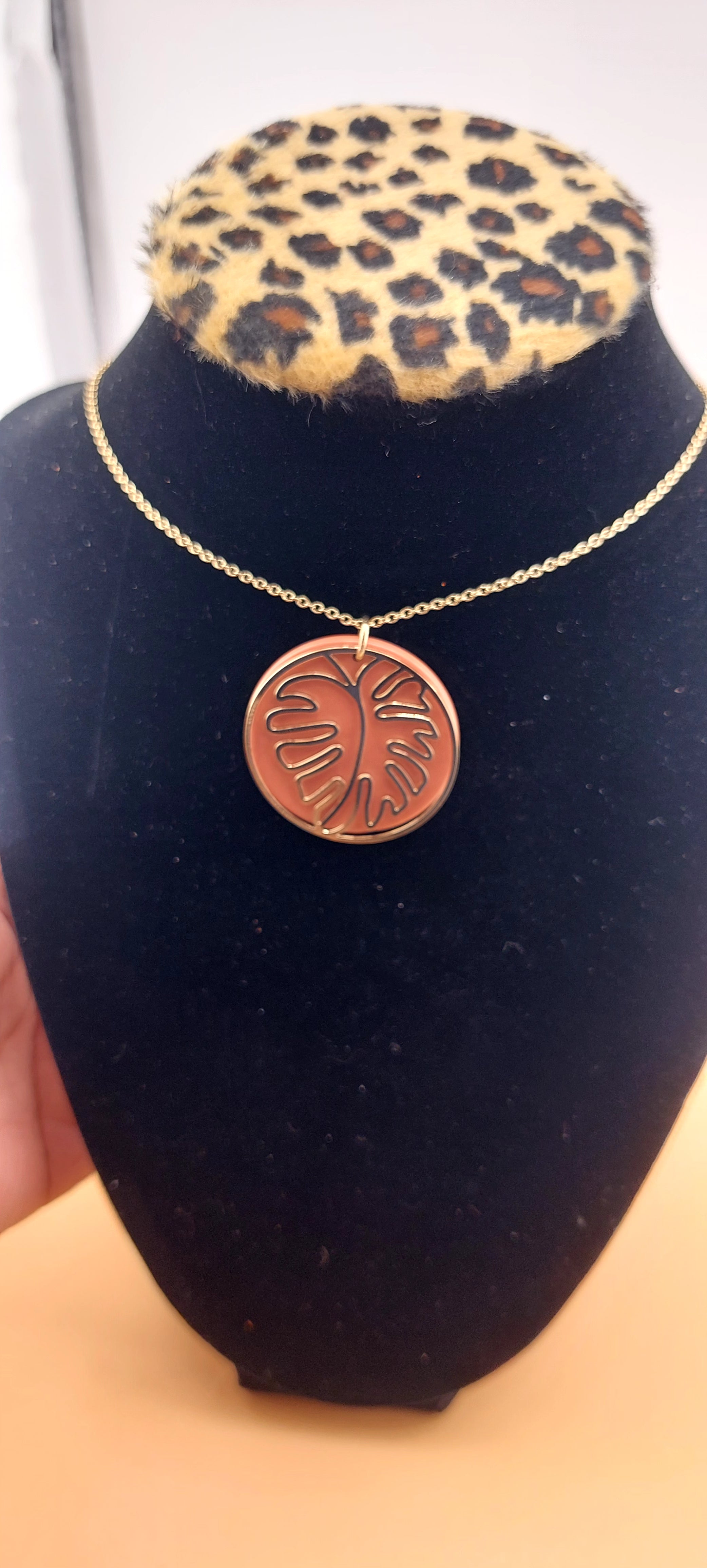 A gold-colored leaf-shaped necklace displayed on a black mount with a leopard print pattern.