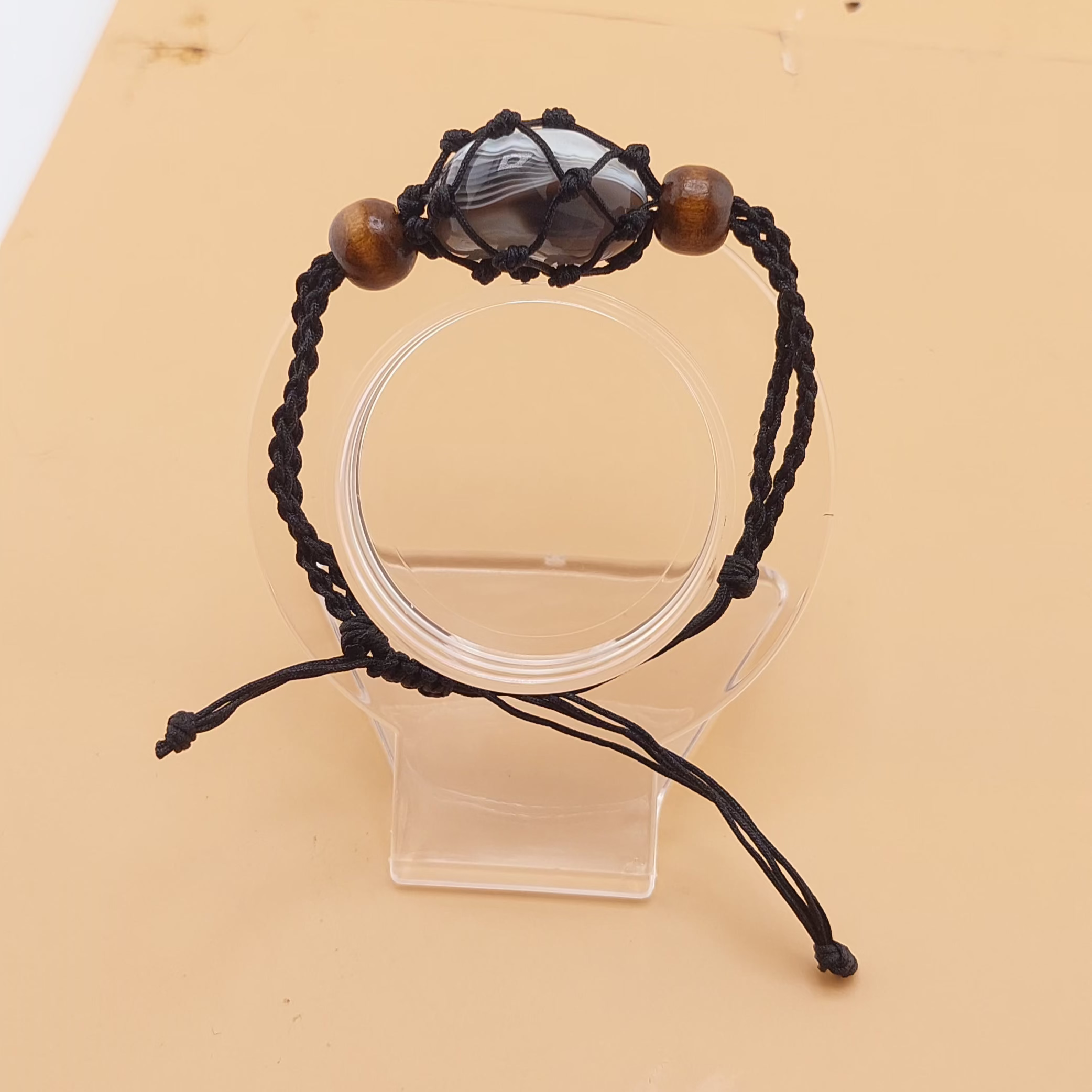 A black Macrame tumbled stone bracelet displayed on a clear stand against a beige background.