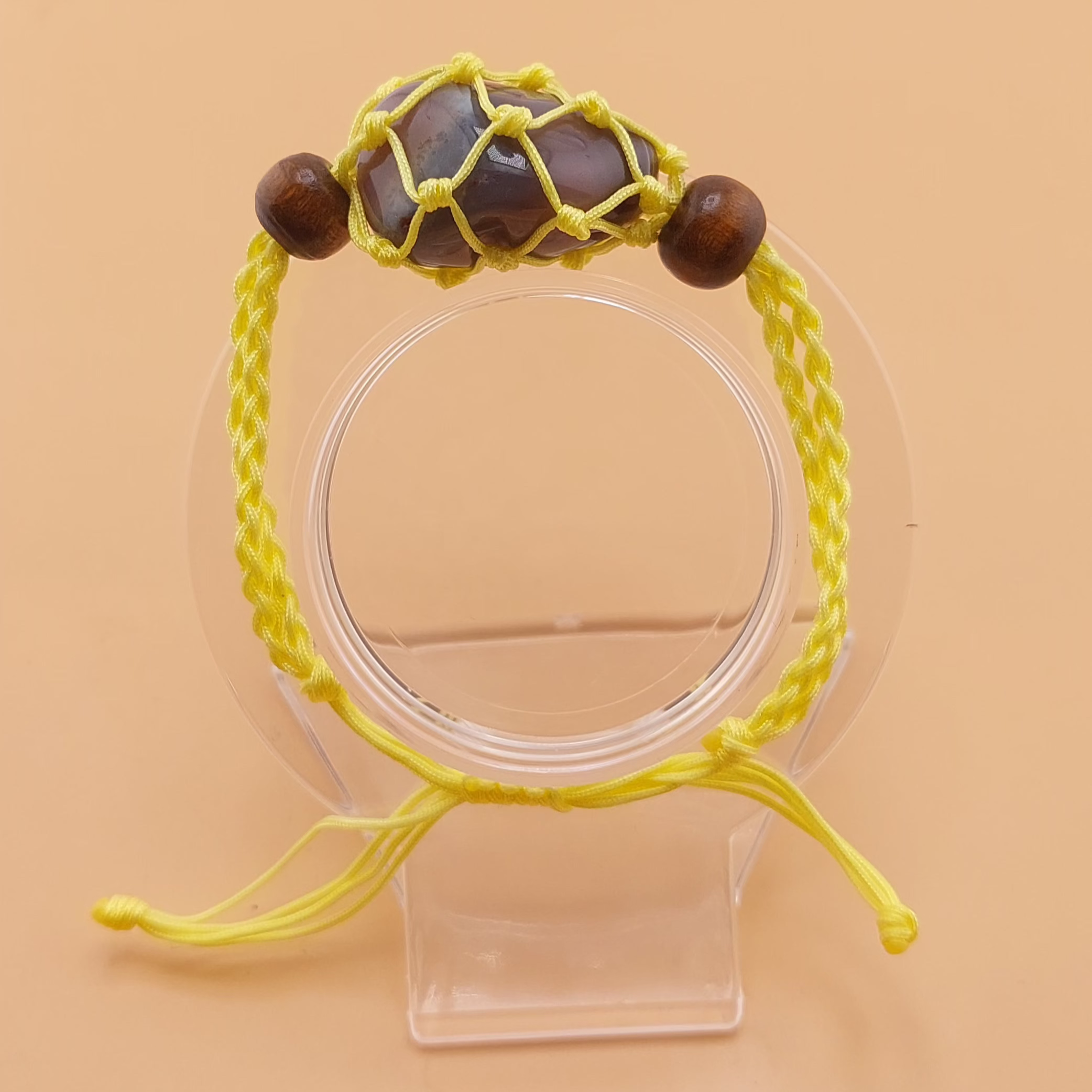 A yellow Macrame tumbled stone bracelet displayed on a clear stand against a beige background.
