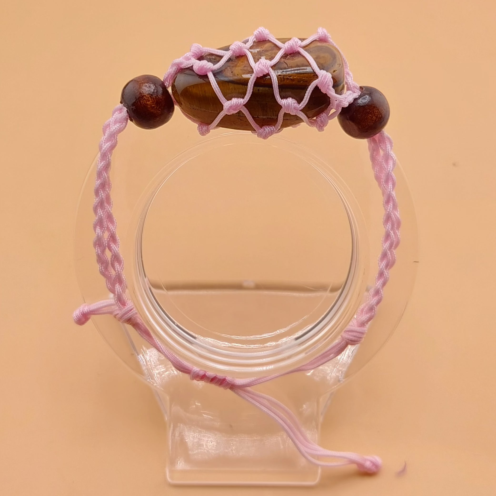A pink Macrame tumbled stone bracelet displayed on a clear stand against a beige background.