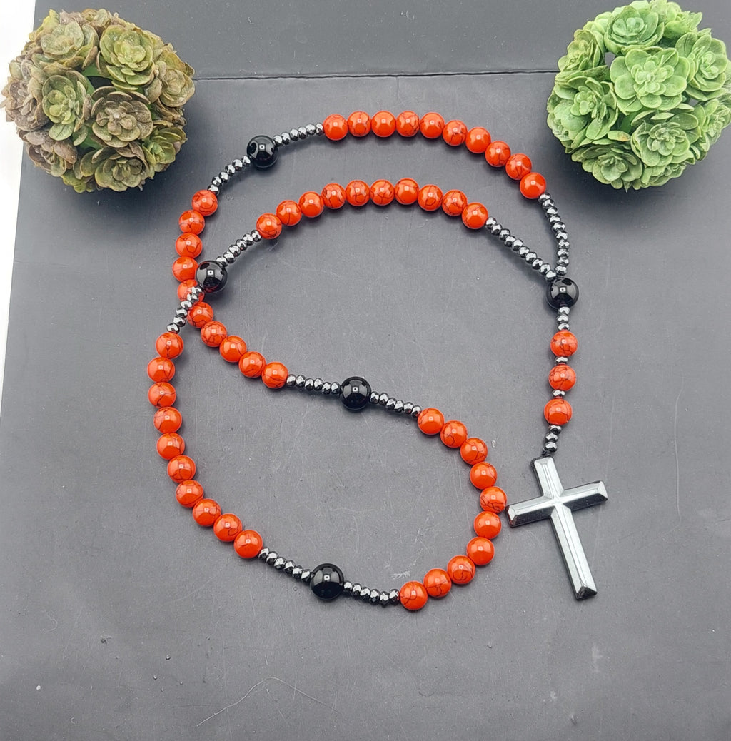 A men's rosary necklace with orange beads and black accents, featuring a silver cross pendant, displayed against a dark background with decorative greenery.