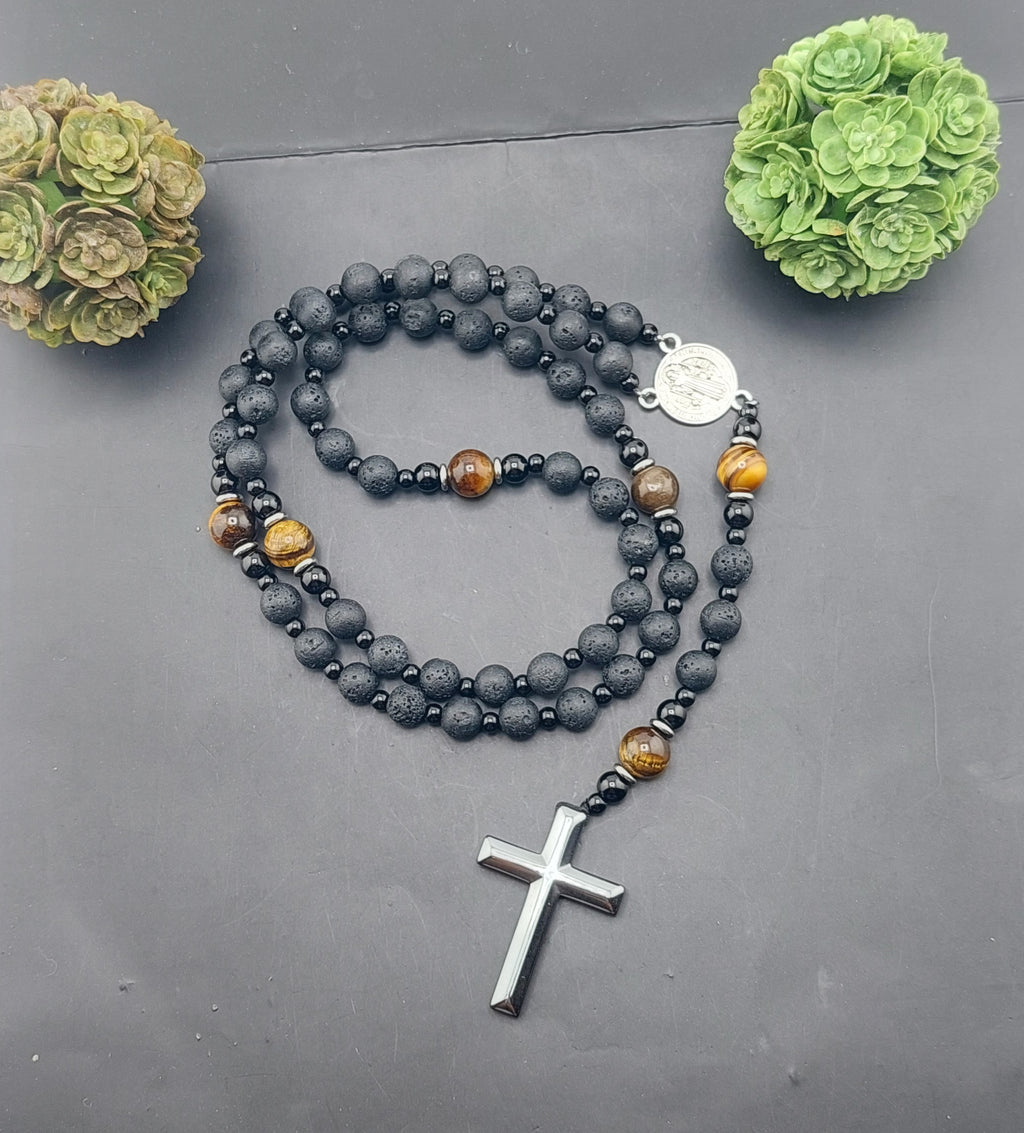 A men's rosary necklace with orange beads and black accents, featuring a silver cross pendant, displayed against a dark background with decorative greenery.