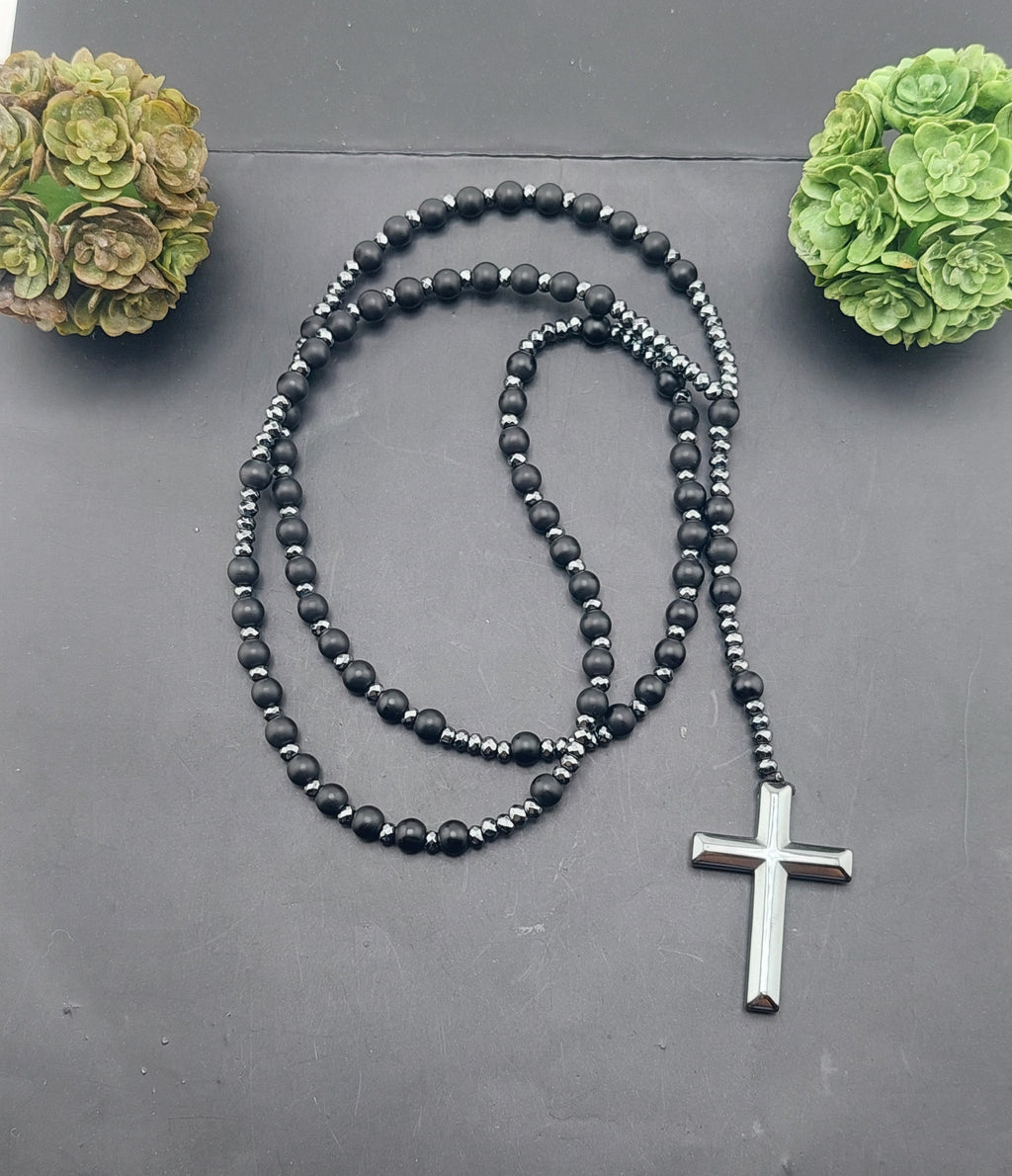 A men's rosary necklace with orange beads and black accents, featuring a silver cross pendant, displayed against a dark background with decorative greenery.