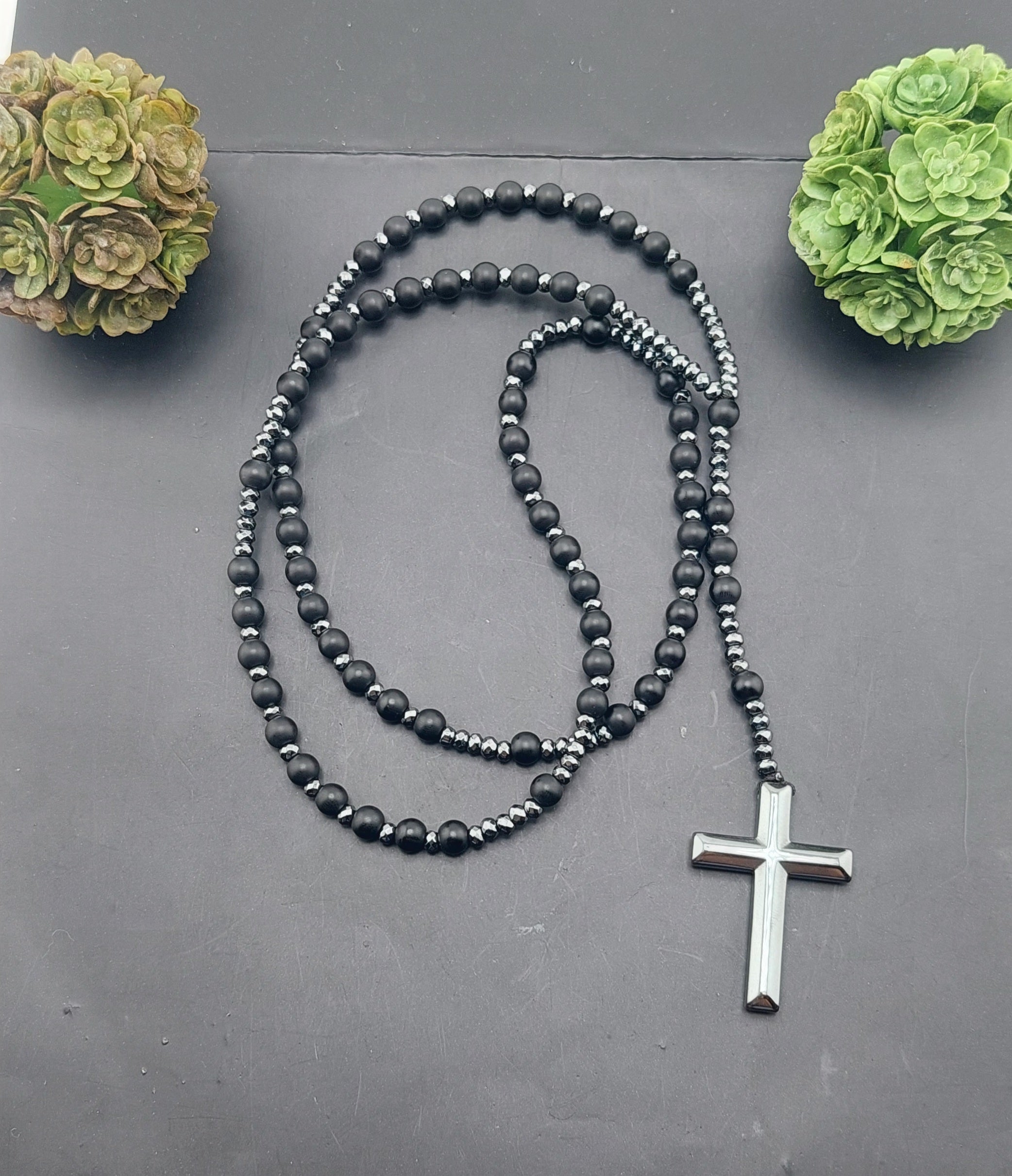A men's rosary necklace with orange beads and black accents, featuring a silver cross pendant, displayed against a dark background with decorative greenery.