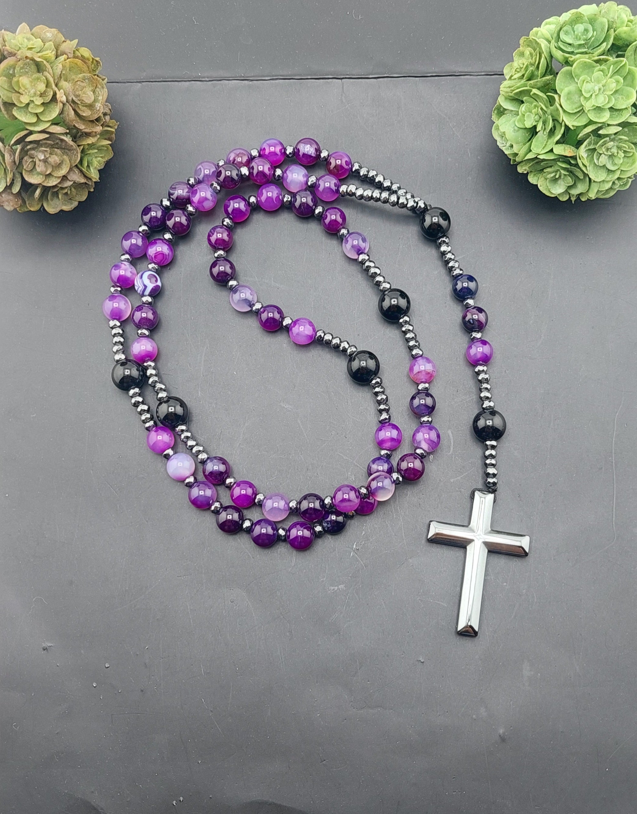 A men's rosary necklace with orange beads and black accents, featuring a silver cross pendant, displayed against a dark background with decorative greenery.