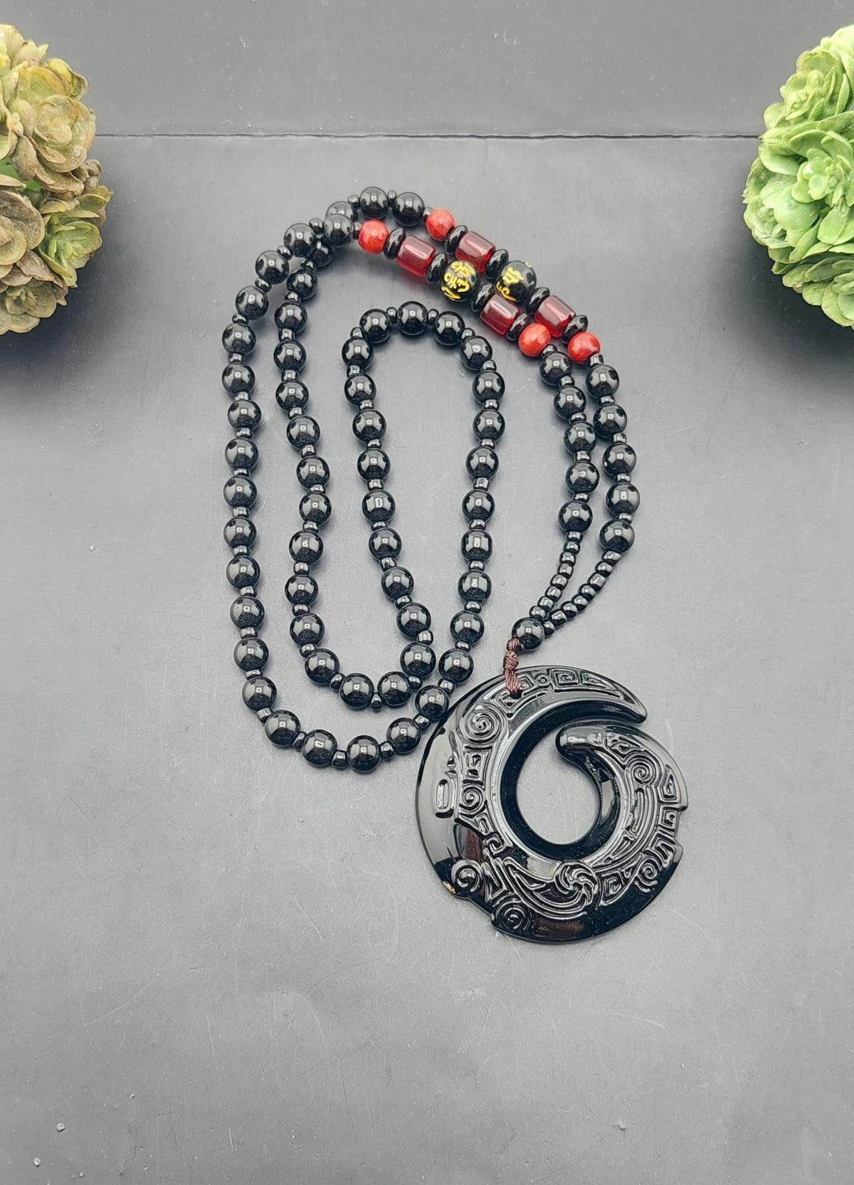 A black obsidian necklace with a beaded chain and a large black pendant featuring an intricate design, displayed against a grey background with decorative flowers on either side.