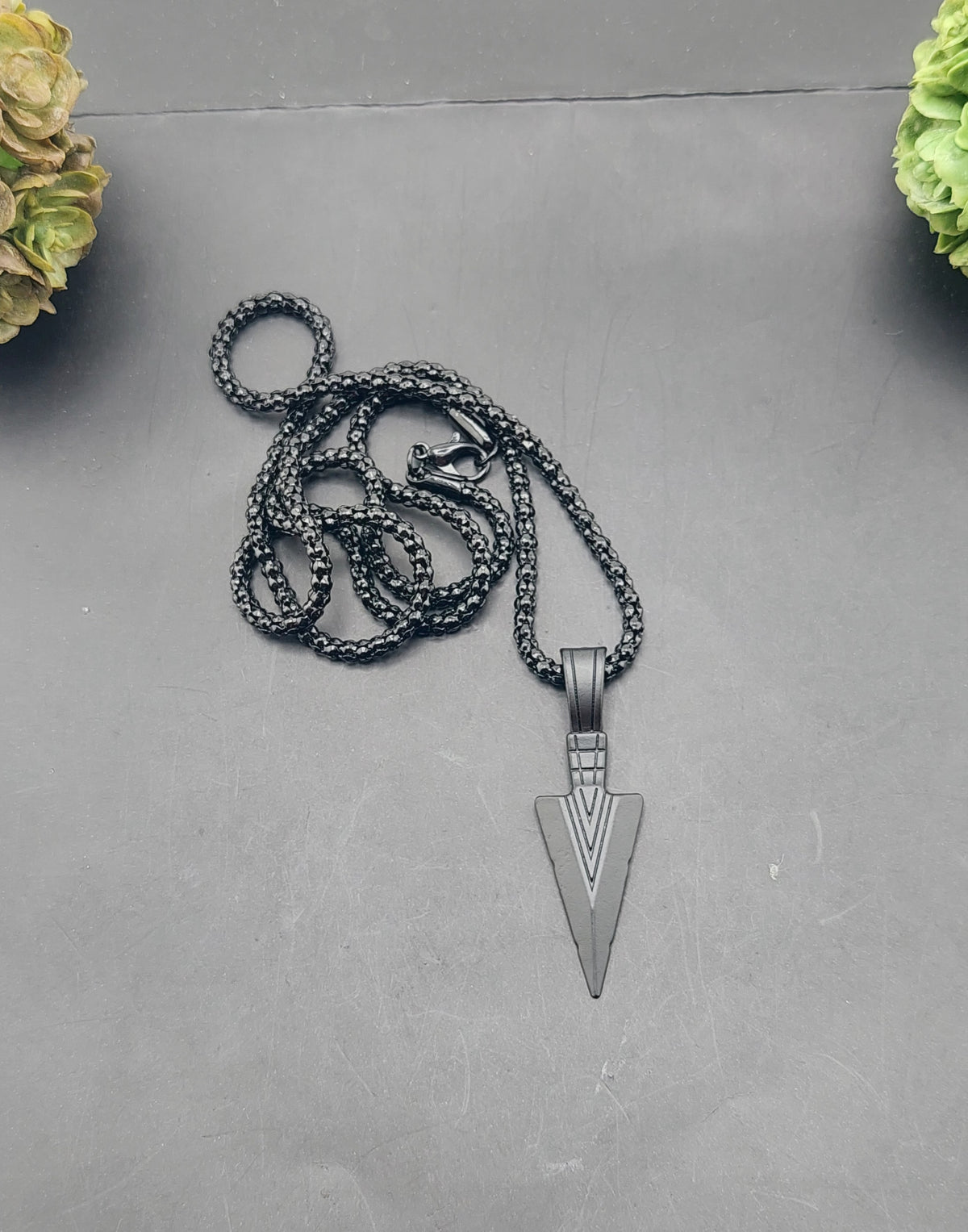 A black spear head necklace displayed against a neutral background with two yellow flowers on either side.