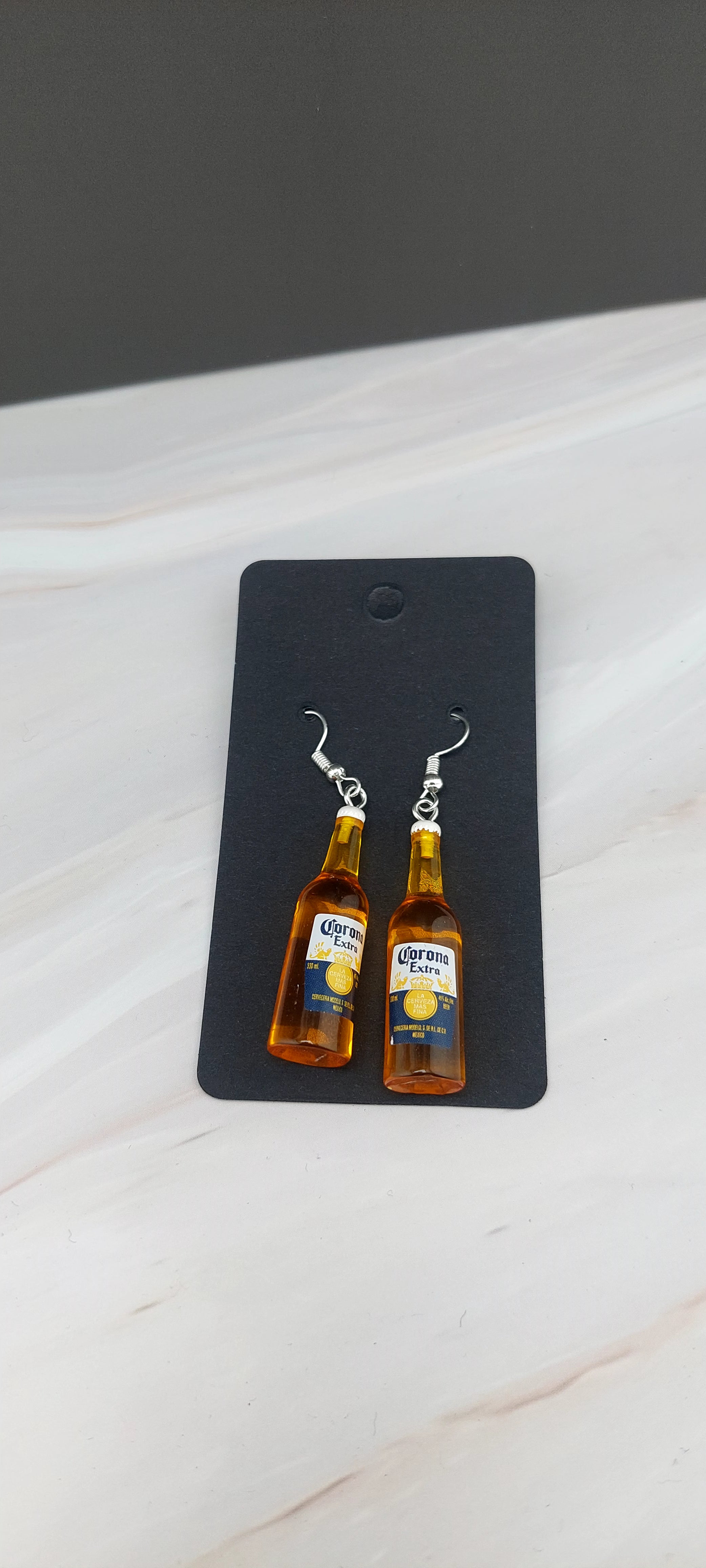 A pair of earrings designed to resemble Corona extra bottles, displayed on a black holder against a marble background.
