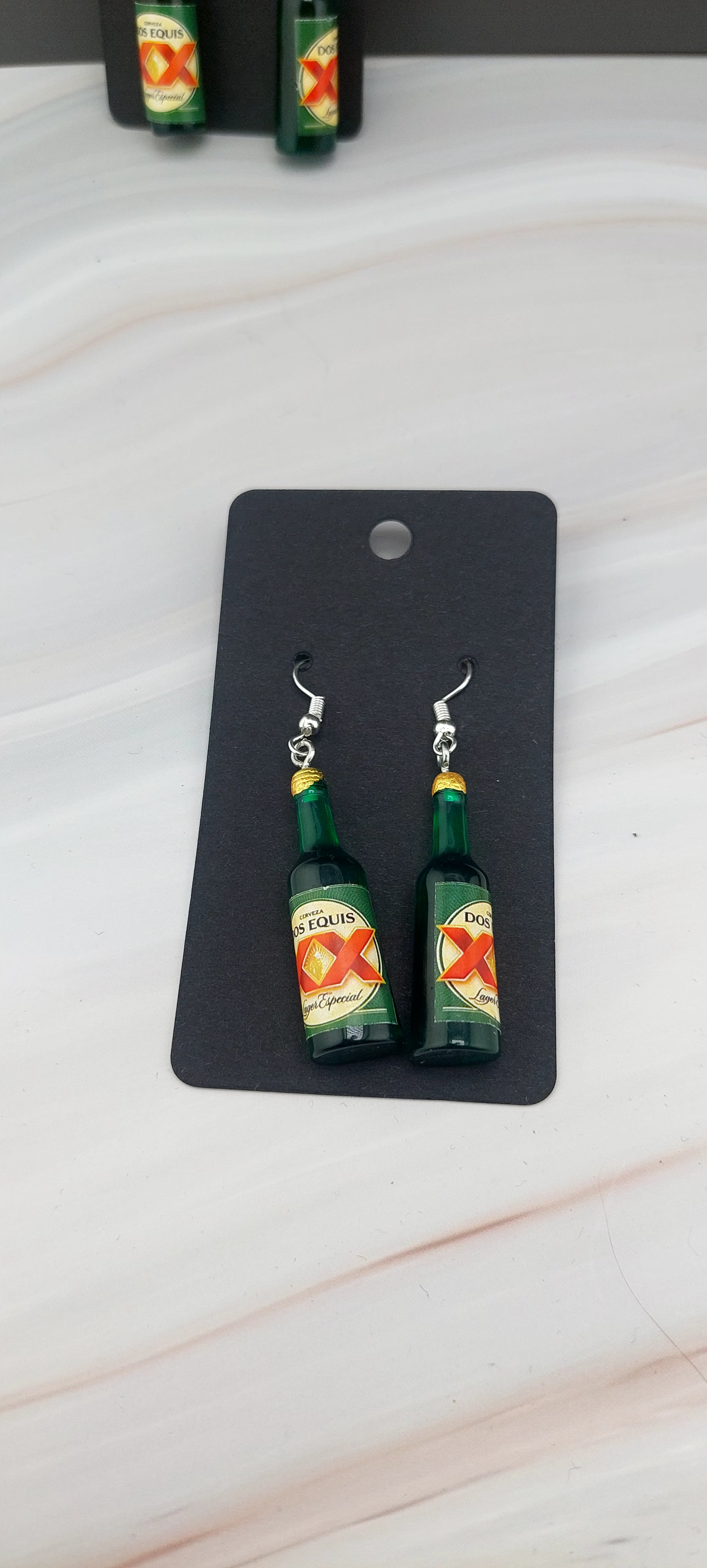 A pair of Dos Equis bottle-shaped earrings displayed on an earrings stand against a marble background.