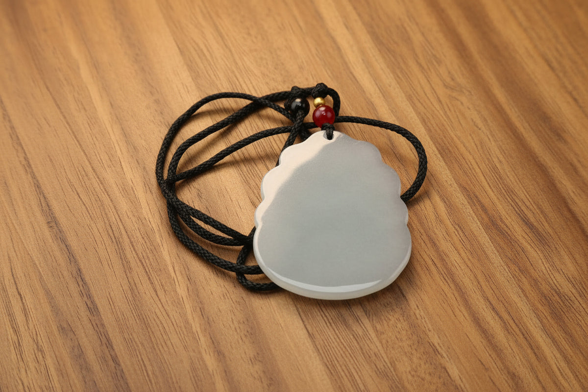 A carved jadeite pendant with a scalloped edge, featuring a milky white and light green coloration, suspended from a black braided cord with a small red bead accent, set against a dark gray background.