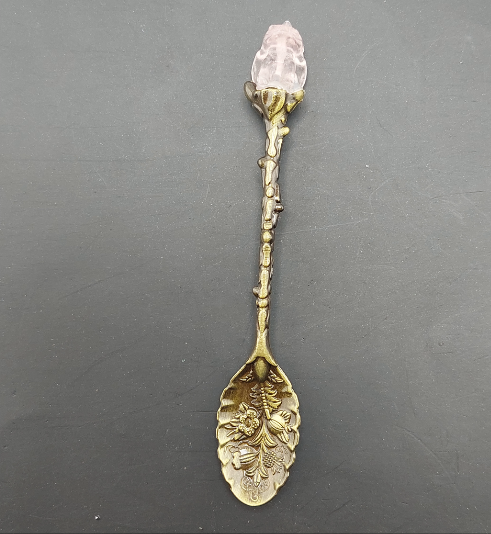 A gold-colored metal tea spoon with a floral design and a pink flower-shaped handle tip, against a dark background.