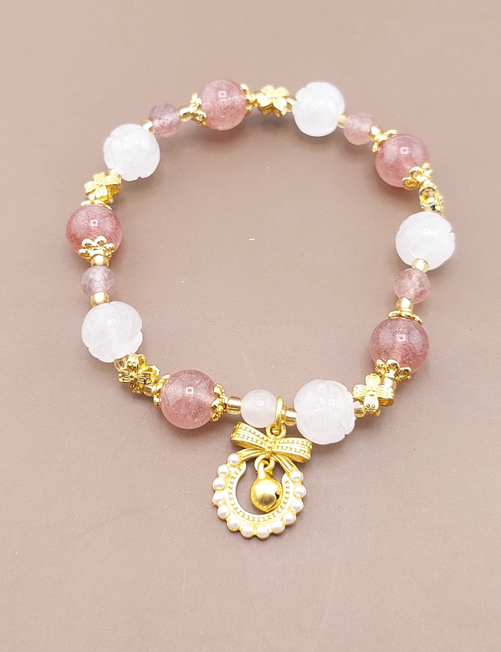 A gold-colored metal bracelet with white and pink beaded details, featuring a gold bow charm and a bell-shaped charm.