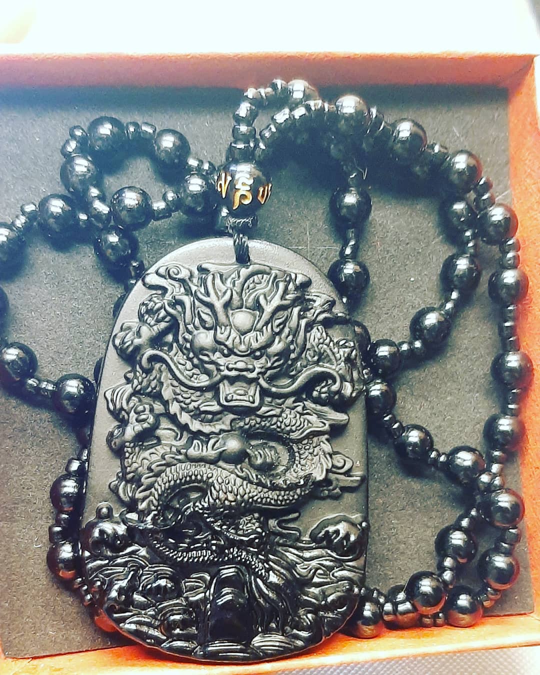 A black obsidian dragon carved necklace presented on a box, featuring a detailed dragon design against a beaded chain background.