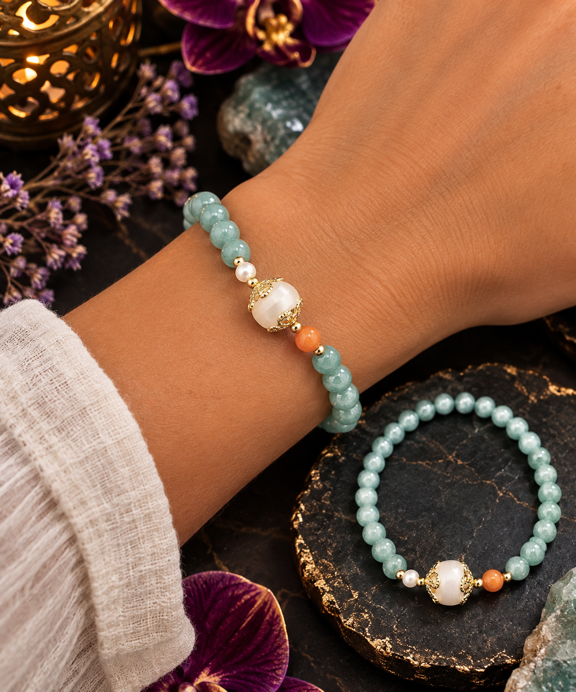 A close-up of a person's wrist wearing a beaded stretch bracelet made of light aqua beads with a white pearl-like center bead, gold-toned spacer, and small orange beads. Another matching bracelet is displayed on a dark stone slab in a styled setting with flowers and crystals.