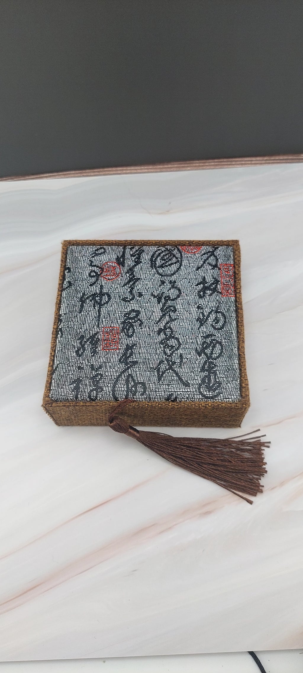 A beige Chinese-style jewelry box with a decorative pattern on the lid, a tassel hanging from the handle, and a white marble background.