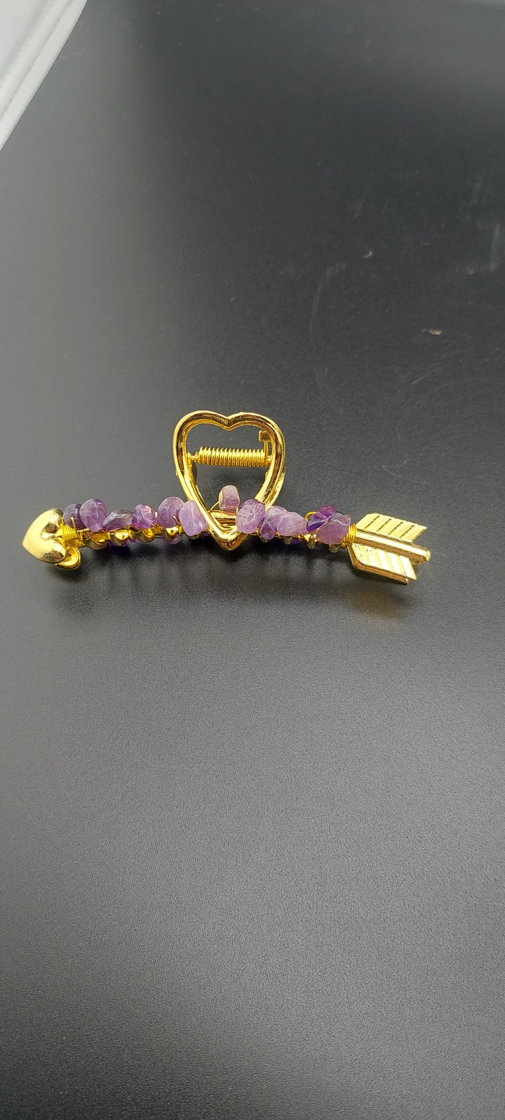 A gold-colored hair clip with a heart and arrow design, featuring purple stones.