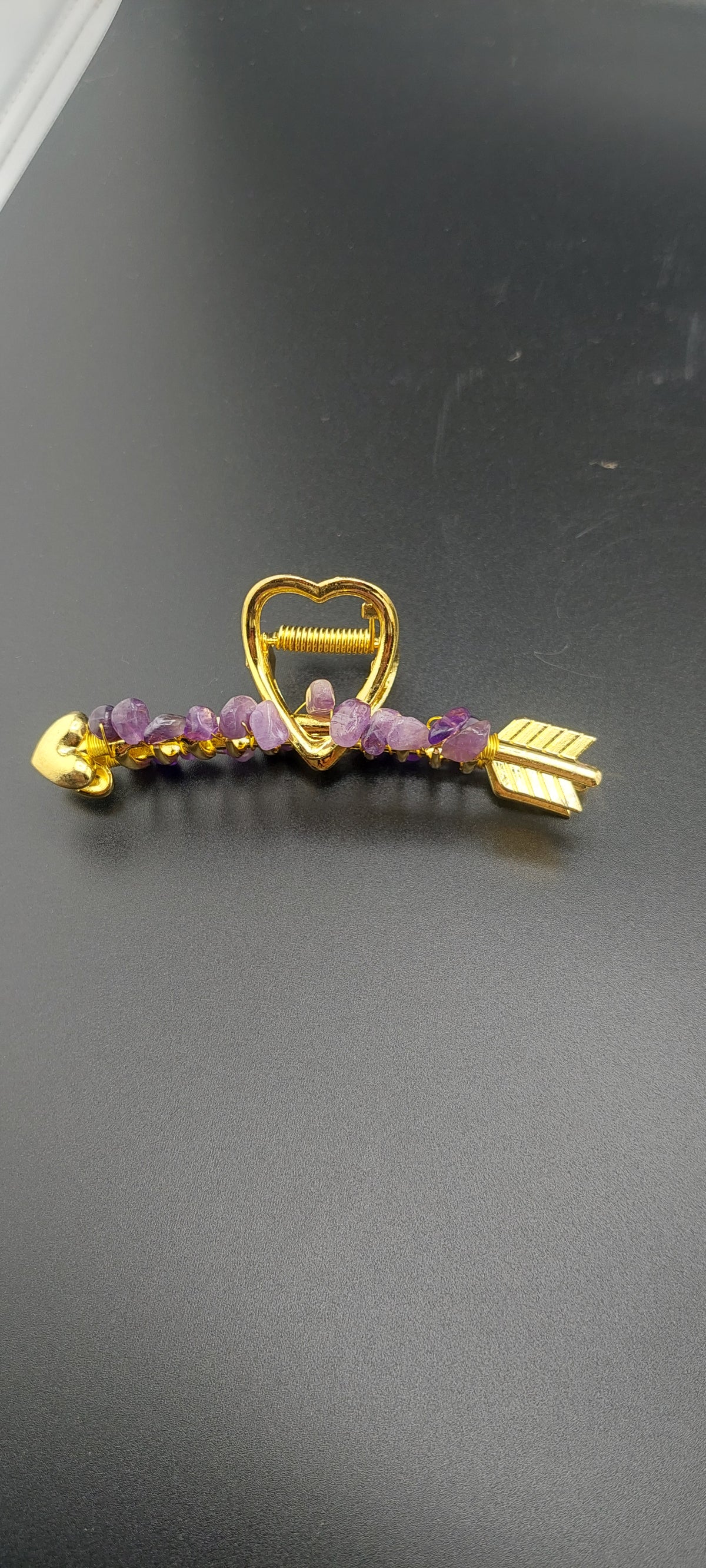 A gold-colored hair clip with a heart and arrow design, featuring purple stones.