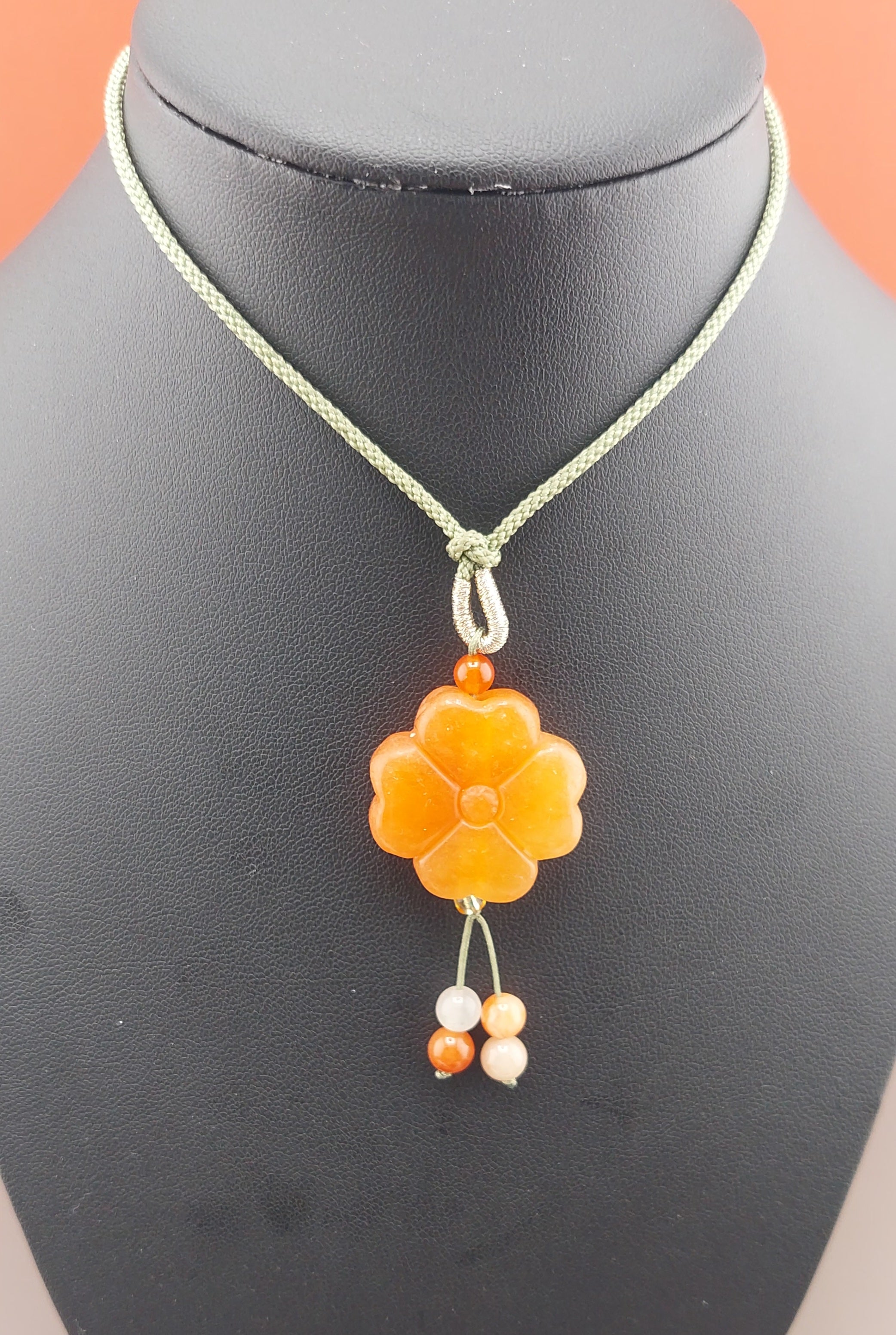 An orange flower necklace displayed on a black mannequin, featuring a floral design with a central orange pendant and small white beads on a gold-tone chain.