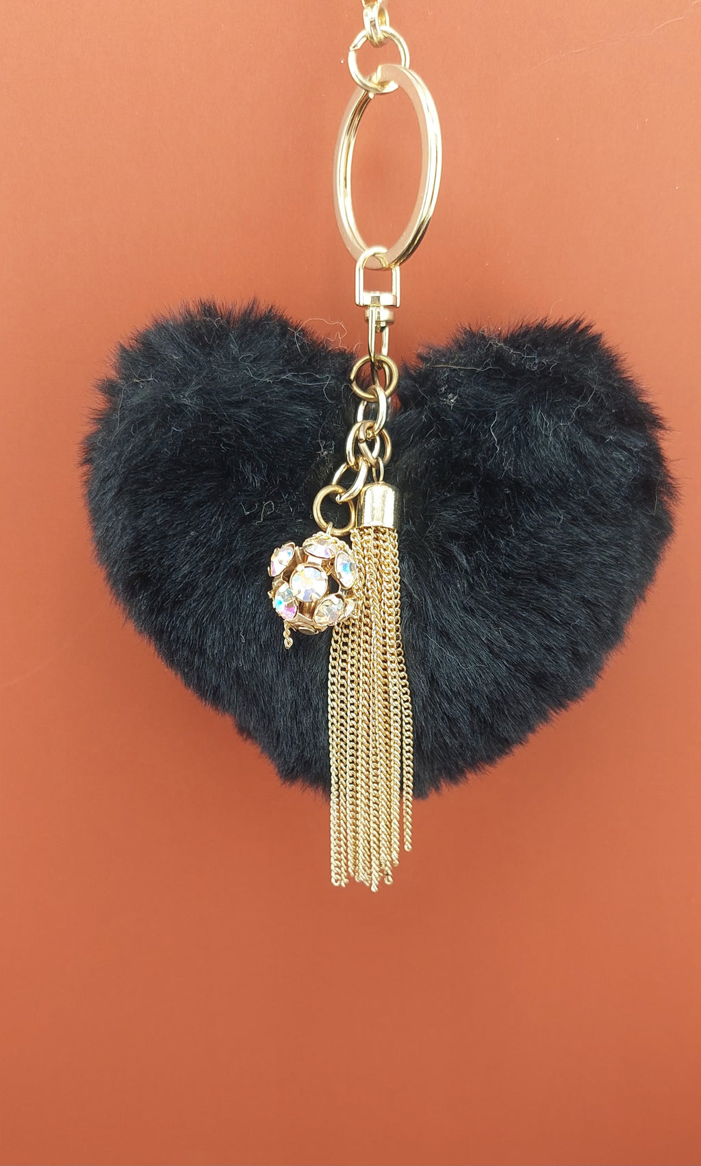 Heart-shaped black fuzzy keychain with gold tassel and charm on a brown background