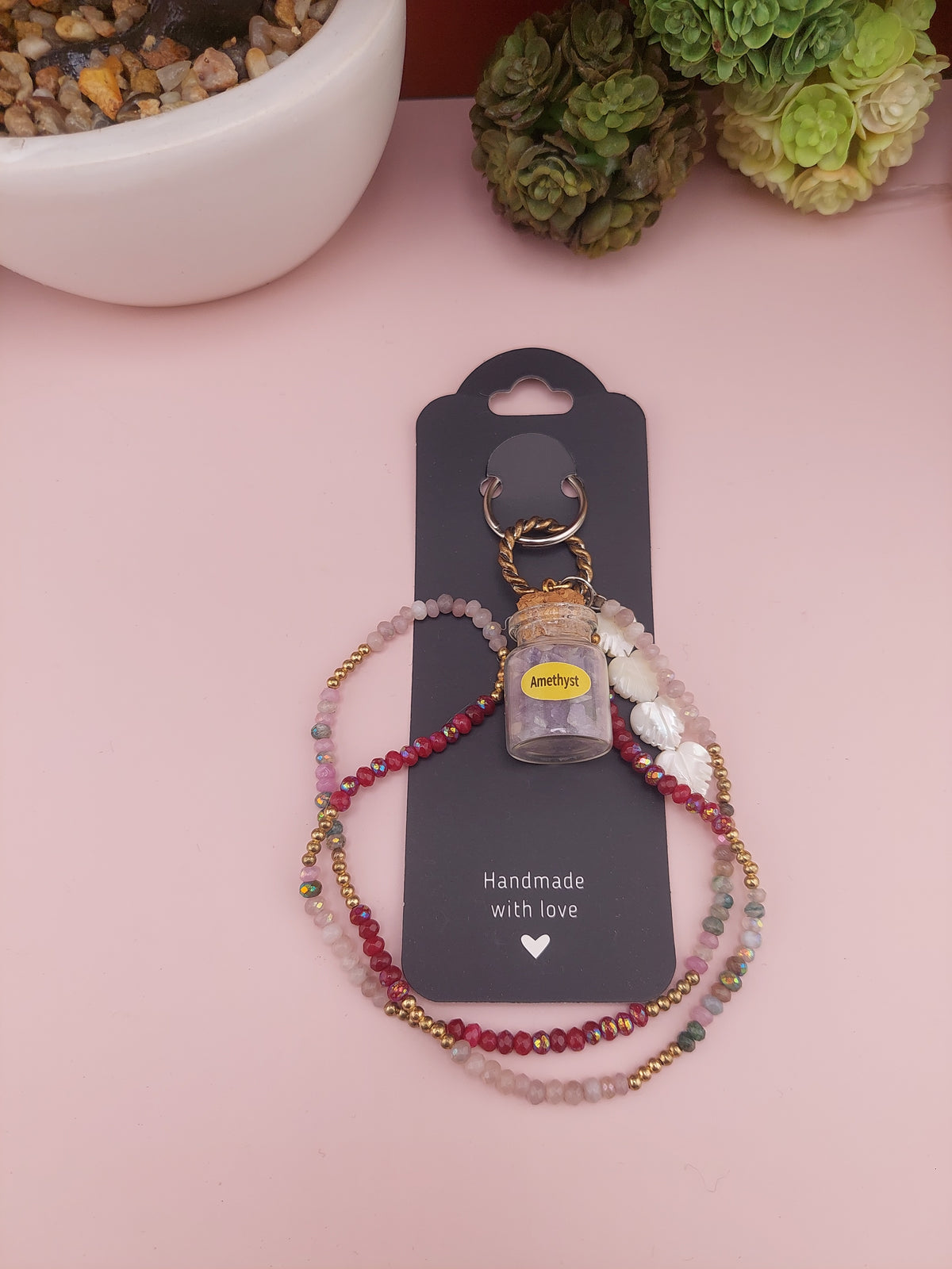 Two beaded bracelets on a black card with 'Handmade with Love' text, placed on a pink surface with a plant in the corner.