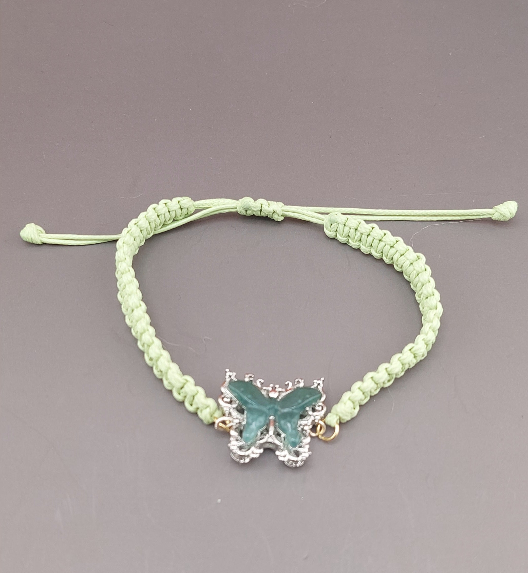 A green macrame anklet with a blue water jade butterfly charm, displayed against a grey background.