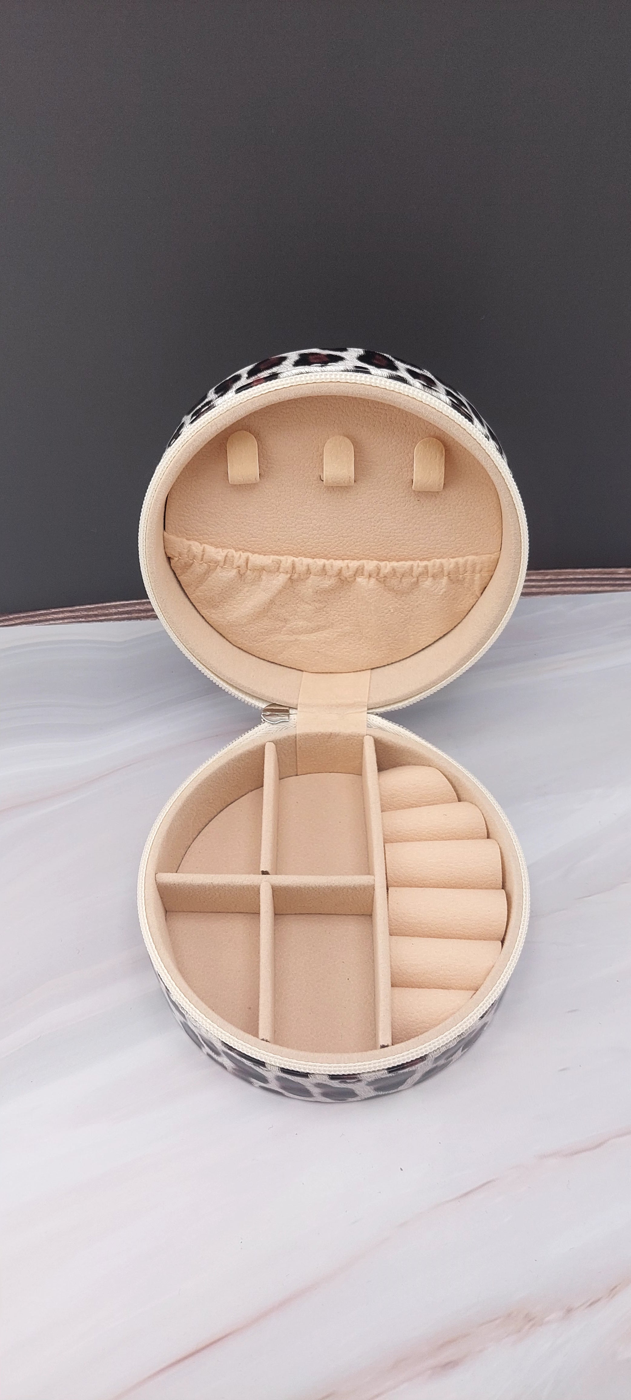 A round jewelry box with a beige interior and patterned exterior, partially open, showing its compartments.