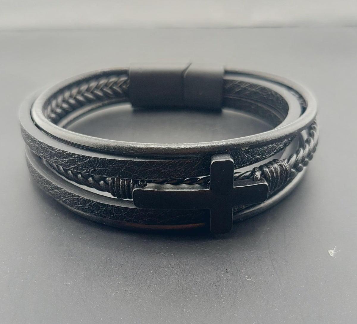 Black leather bracelet with metal cross charm on a gray background