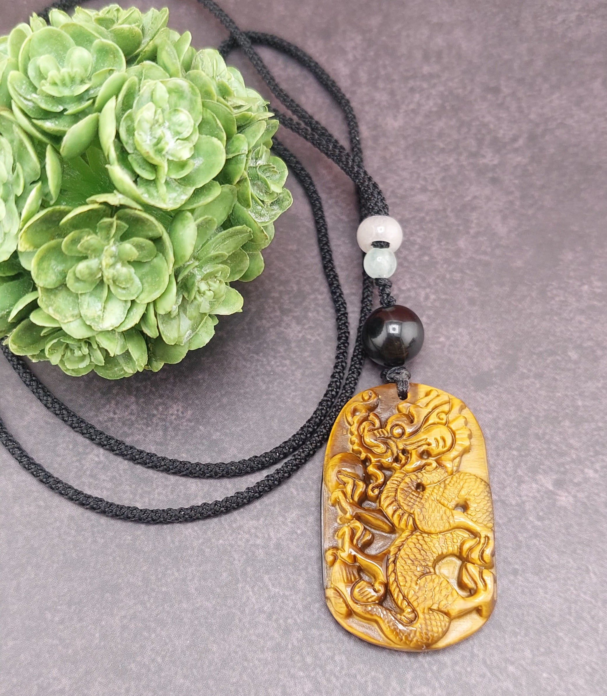 A hand-carved tigers eye dragon pendant on a black cord necklace, displayed next to a green floral decoration.