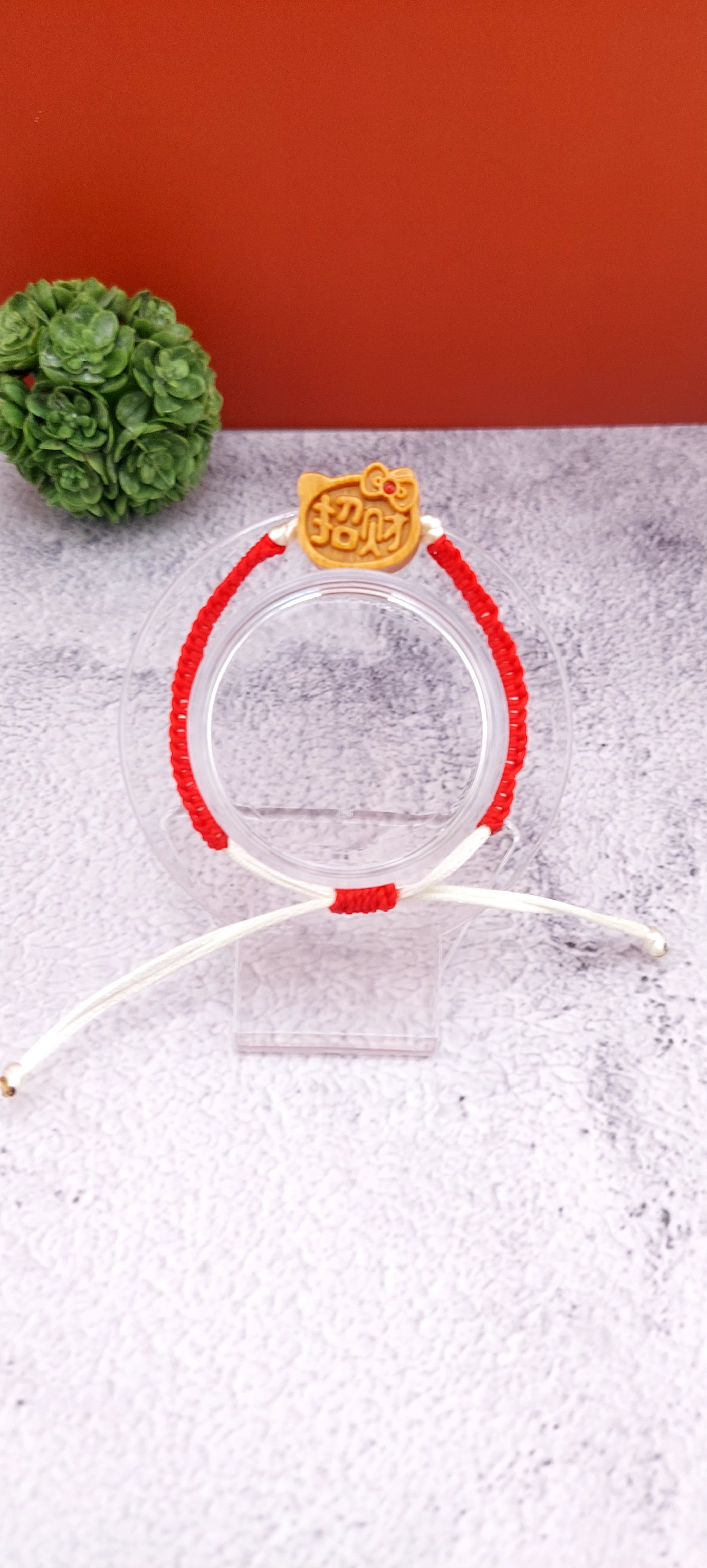 A red and white macrame bracelet with a Hello Kitty charm on a clear display stand against a two-tone background.